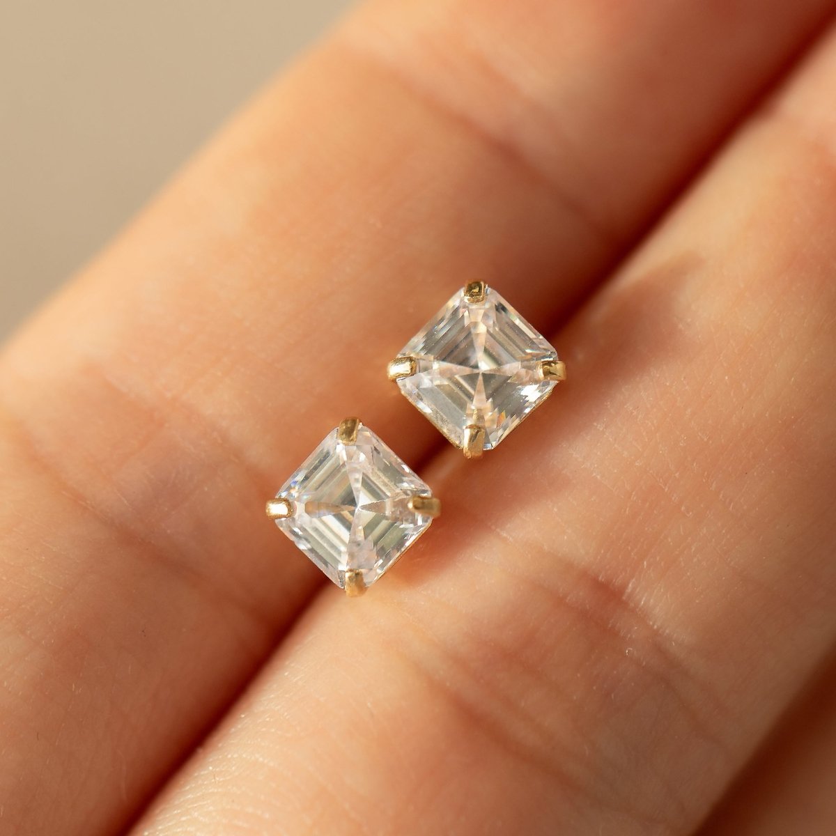 14K gold Asscher-cut white zirconia studs with clear stones, pair held on fingertips