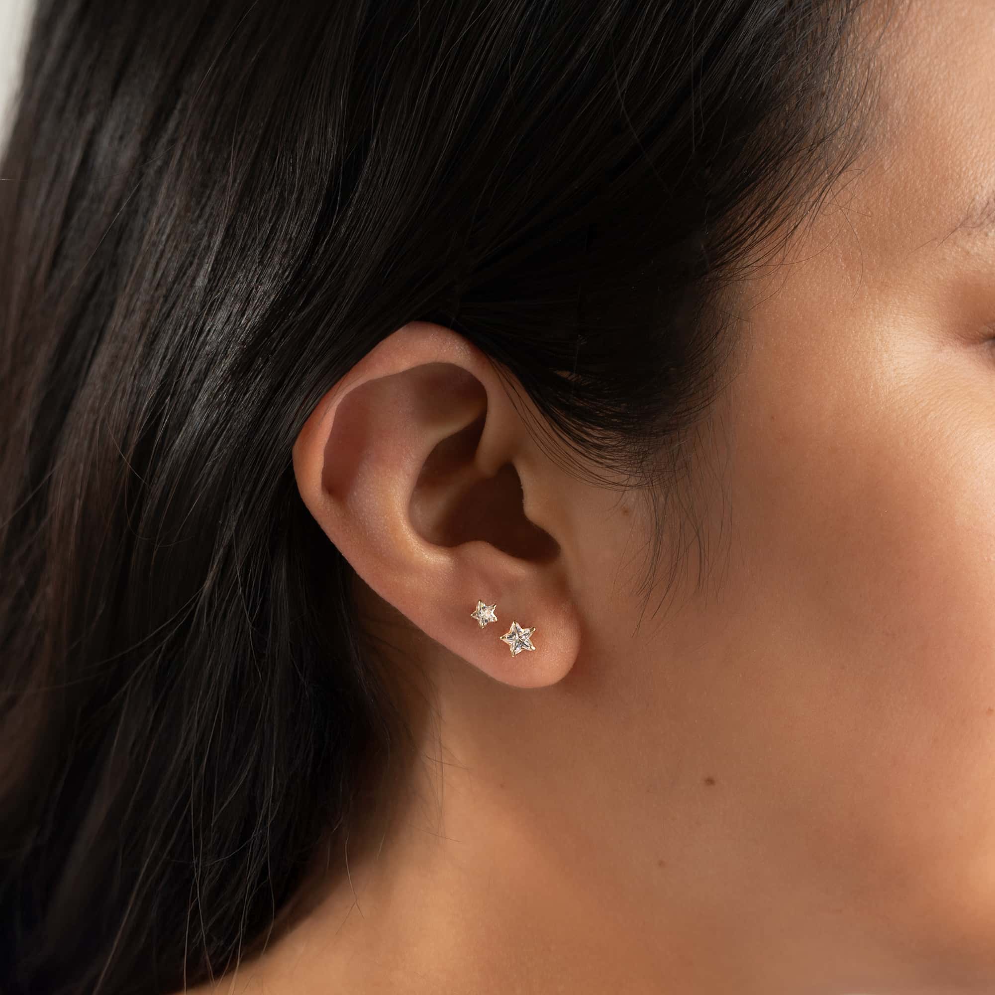 14K gold star studs with clear zirconia stones, shown stacked on a model's lobe