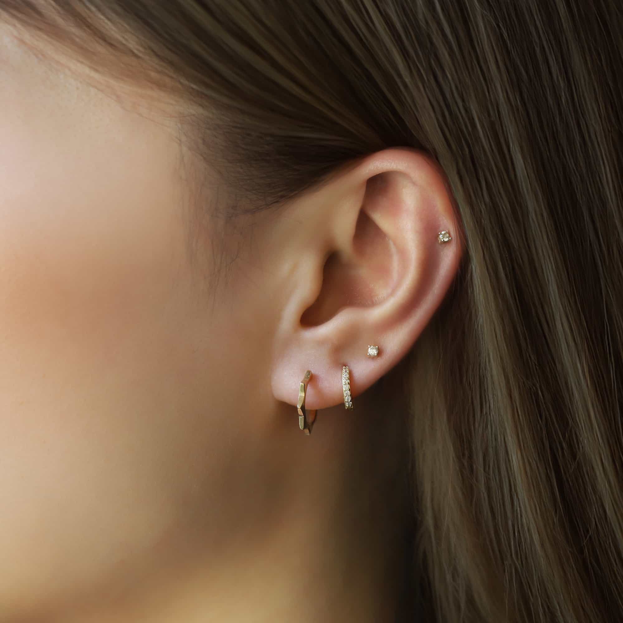 14K gold faceted sunray huggie hoop shown on a model's lobe with stud piercings