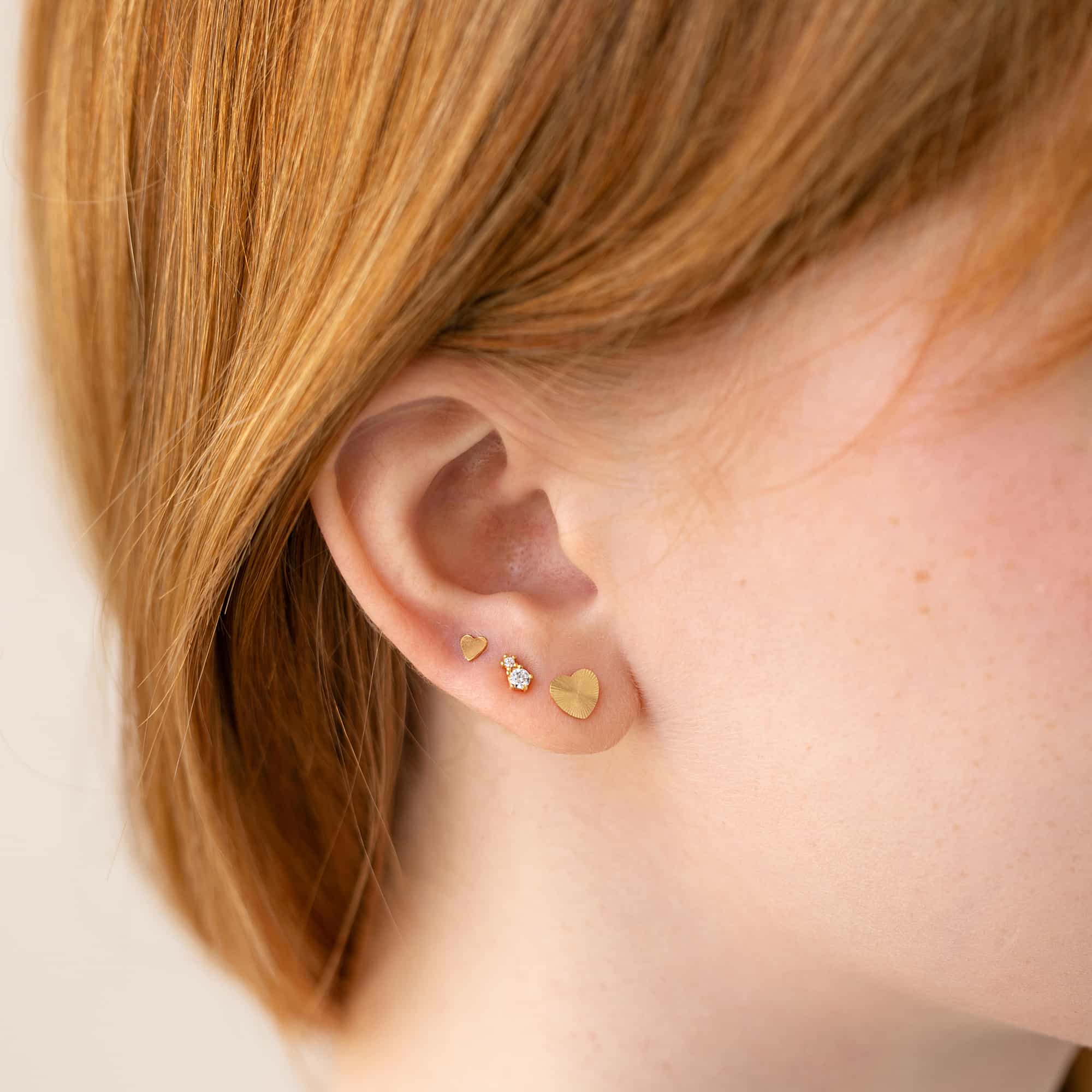 Gold heart studs in a small ear stack shown on a model's lobe