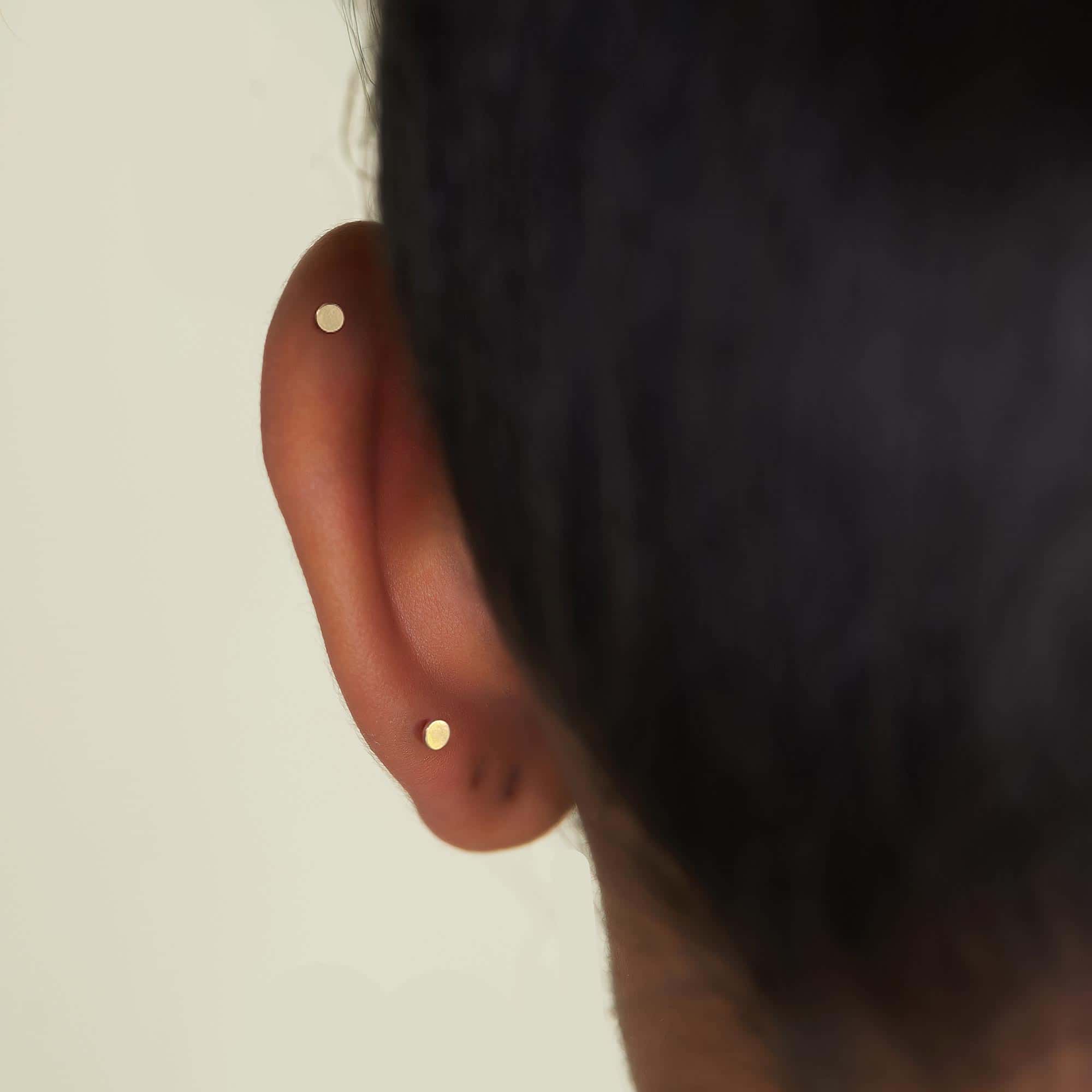 Tiny gold dot flat back studs shown in helix and lobe piercings on a model's ear