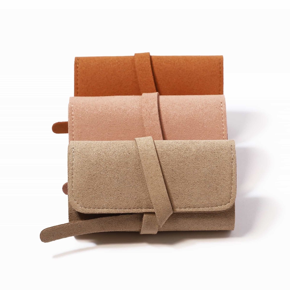 Three travel jewelry rolls in tan, pink, and rust suede stacked, on a white background