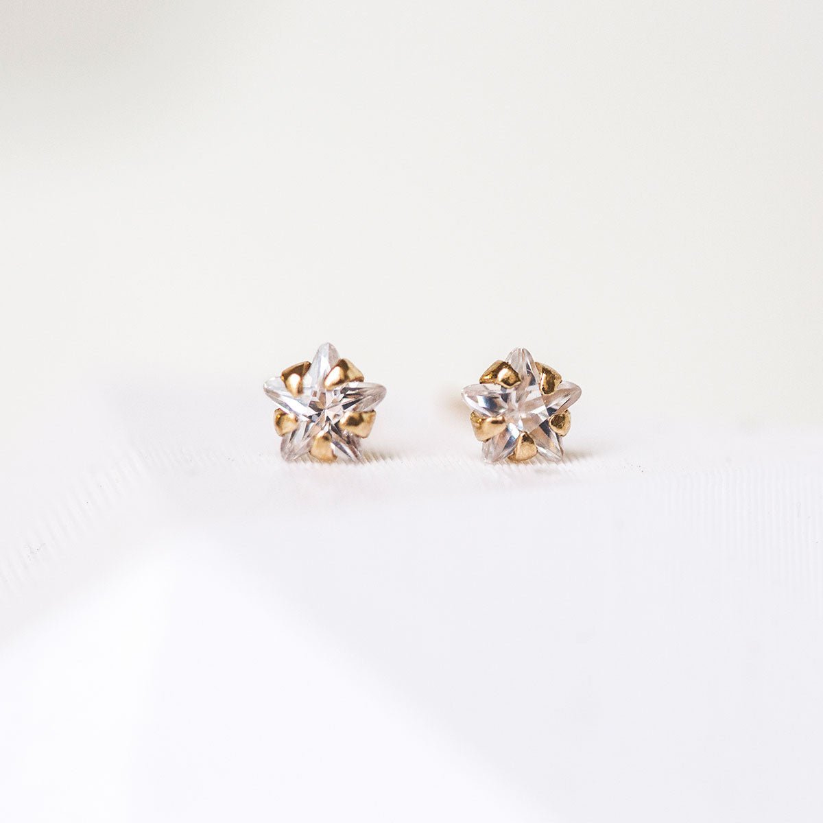 14K gold tiny star stud pair with clear star-shaped stones, on a cream background