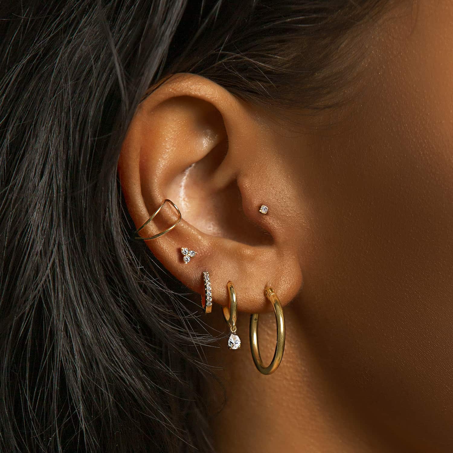 Gold hoops, an ear cuff, a clear-stone drop huggie, and tiny studs styled on a model's ear