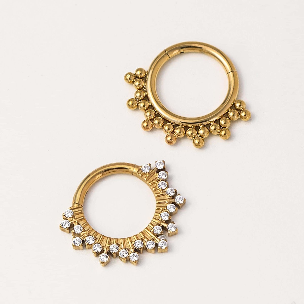 Two gold clicker hoops, one beaded and one set with clear stones, on a cream background