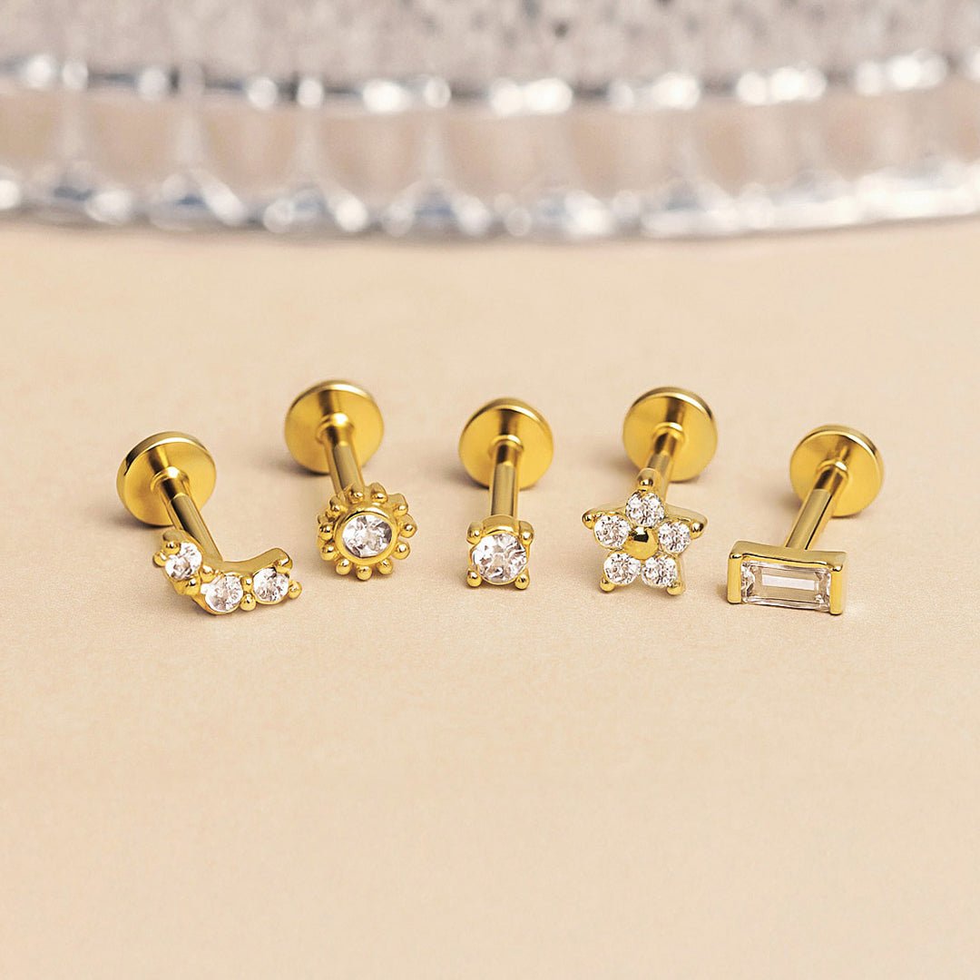 Five gold flat back studs with clear stones in varied shapes including flower, star and bar, on a cream background