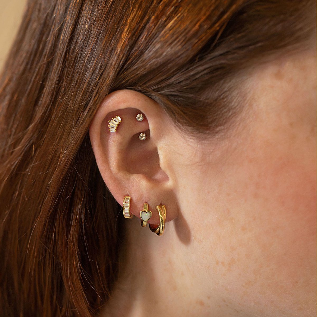 Gold baguette and crystal studs with mini hoops and an opal heart huggie in an ear stack on a model
