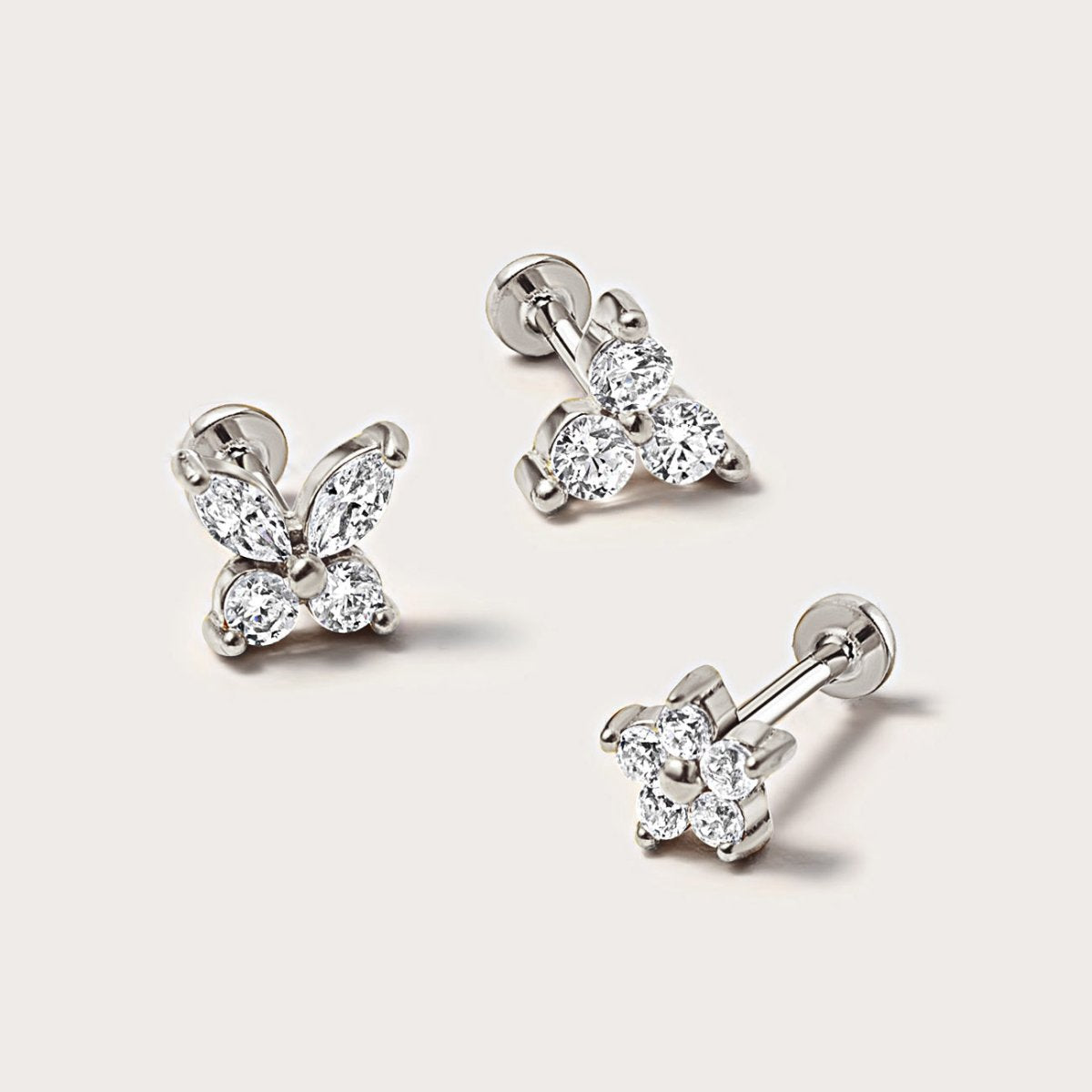 Three silver flat back studs in butterfly, trio and flower styles with clear stones, on a cream background