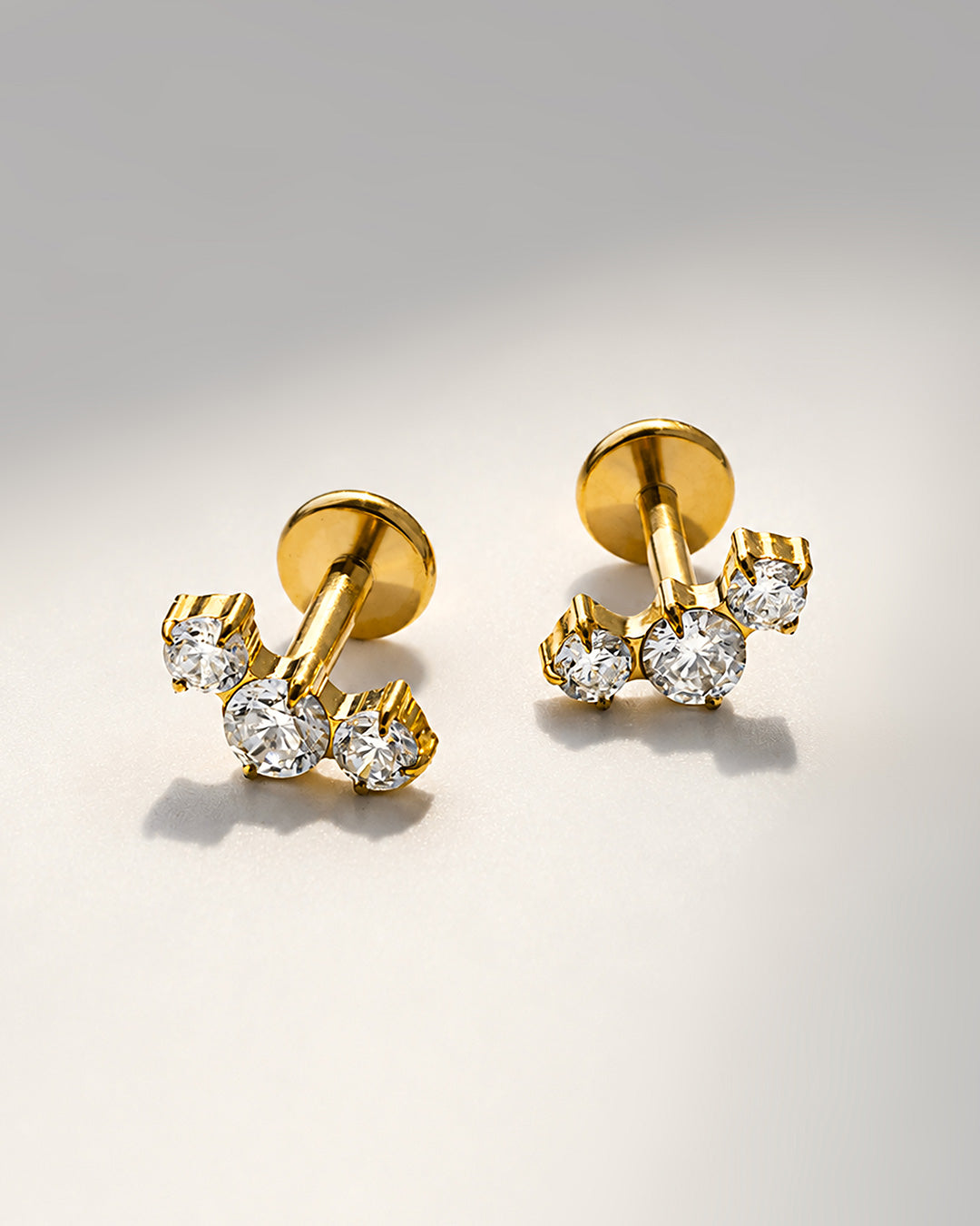 Pair of gold flat back studs set with three round crystals each, resting on a pale surface