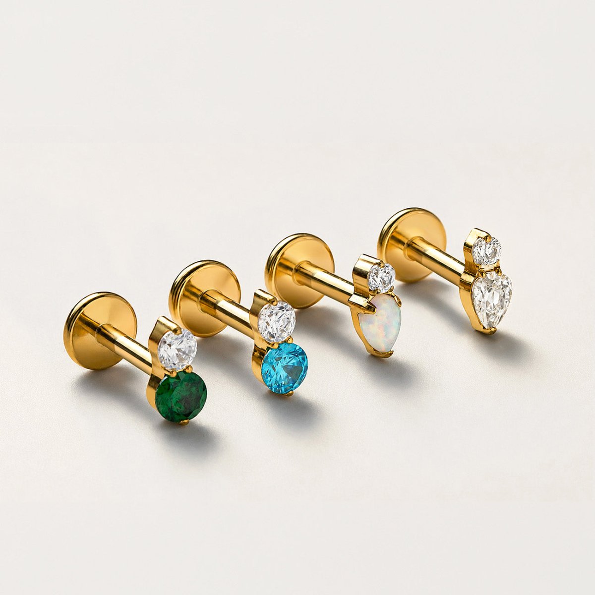 Gold flat back stud earrings with clear and green, blue, white, and pear-shaped stones on a light beige background.
