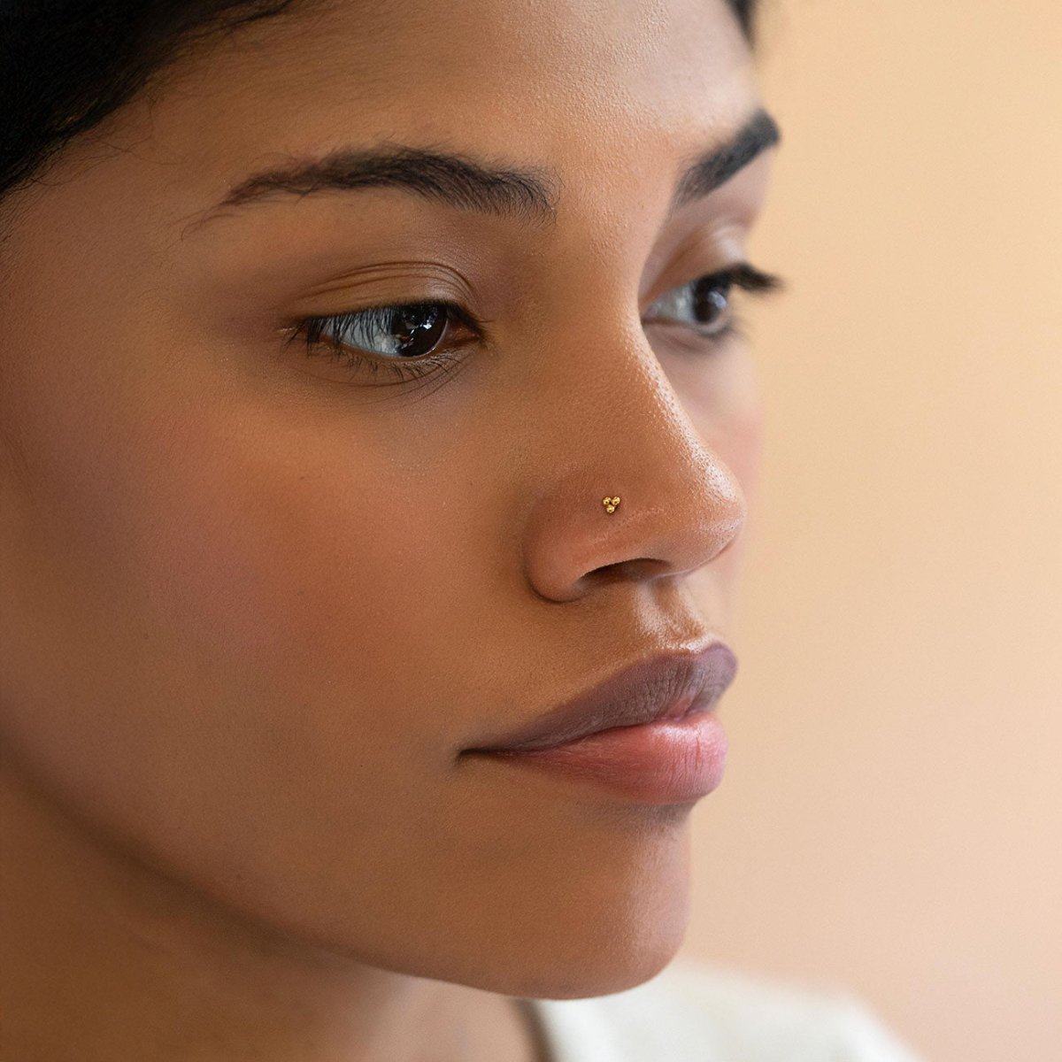 Gold titanium trinity-bead nose stud worn on a model's nostril, soft-focus close-up