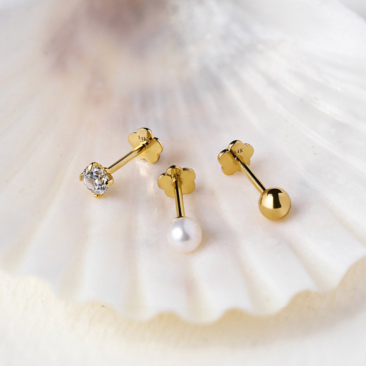 Three gold flat back studs with a clear stone, a pearl and a plain ball, on a cream shell