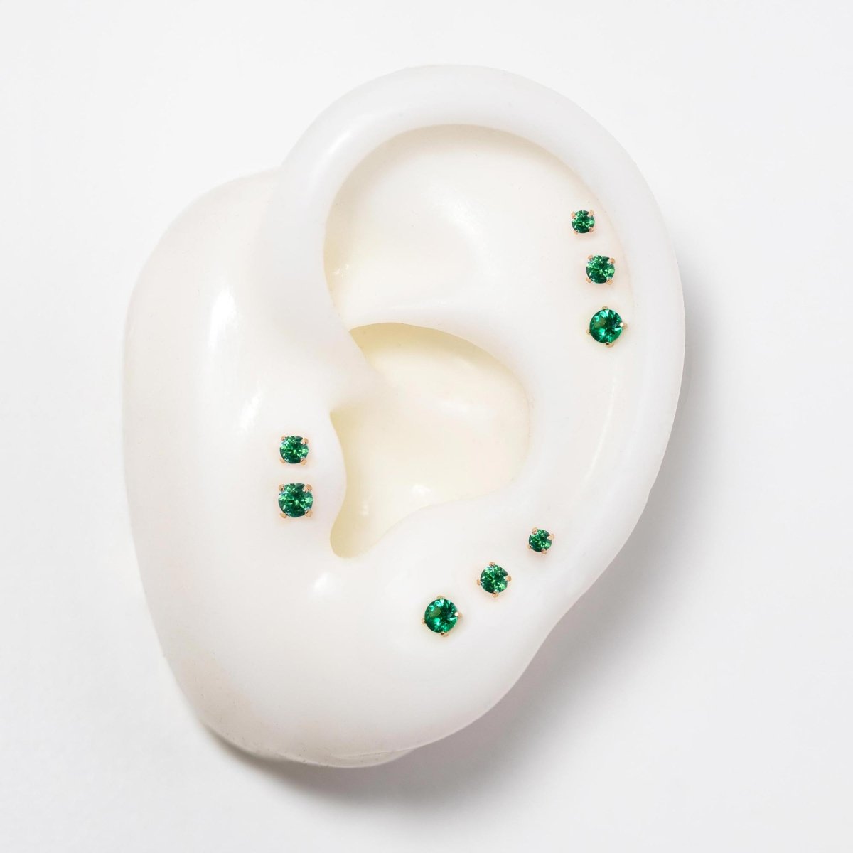 14K gold flat back emerald studs in three sizes on a silicone ear, green stones