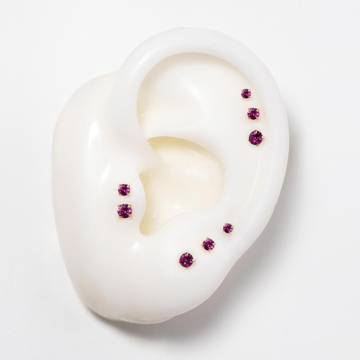 14K gold flat back ruby studs in three sizes on a silicone ear, pinkish-red stones
