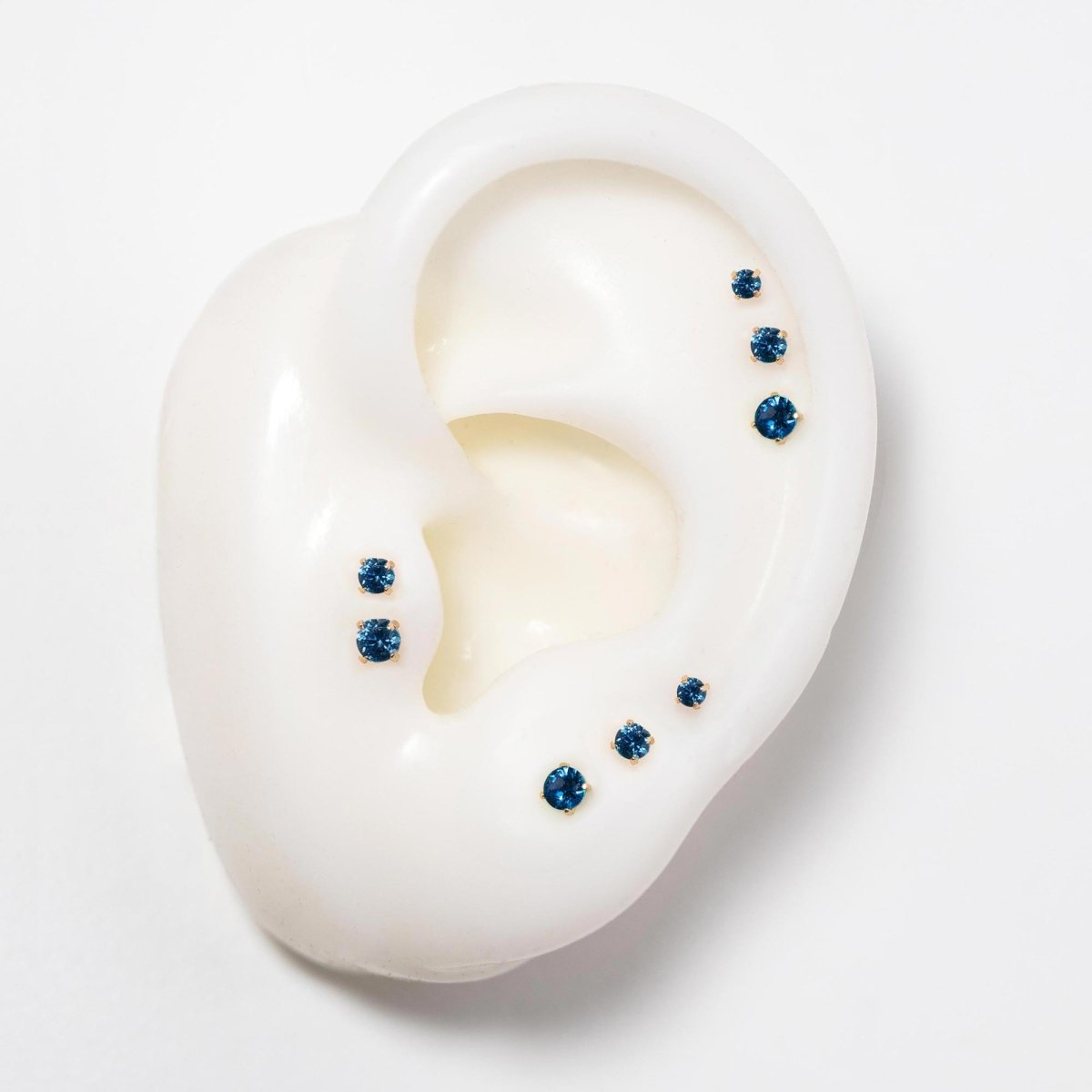 14K gold flat back blue sapphire studs in three sizes on a silicone ear, deep blue stones