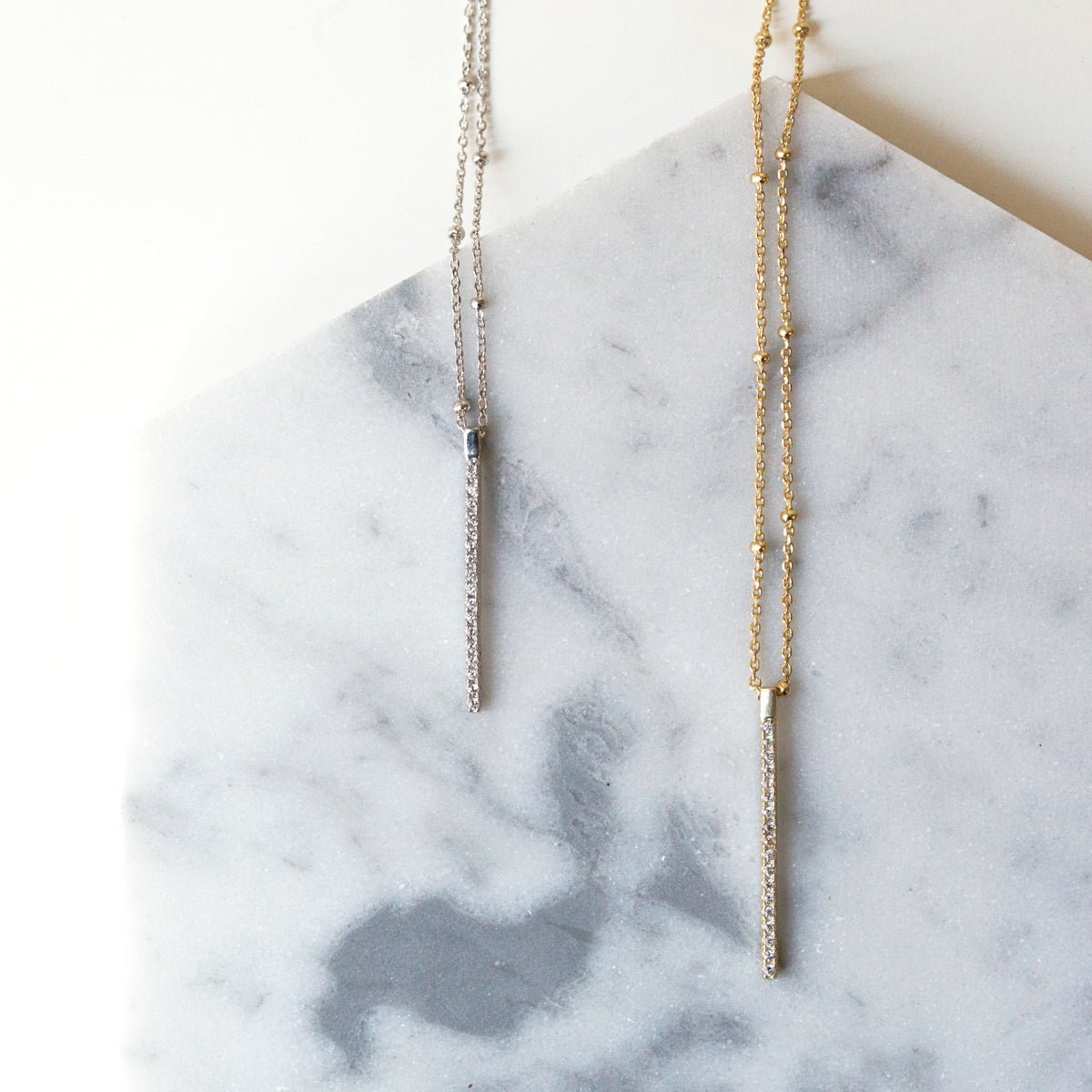 Gold and silver pavé bar pendant necklaces with clear stones, hung against white marble