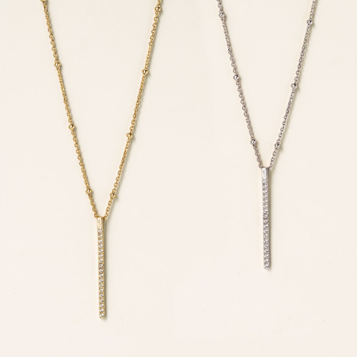 Sterling silver pavé bar pendant on a beaded chain beside a gold version, on a cream background