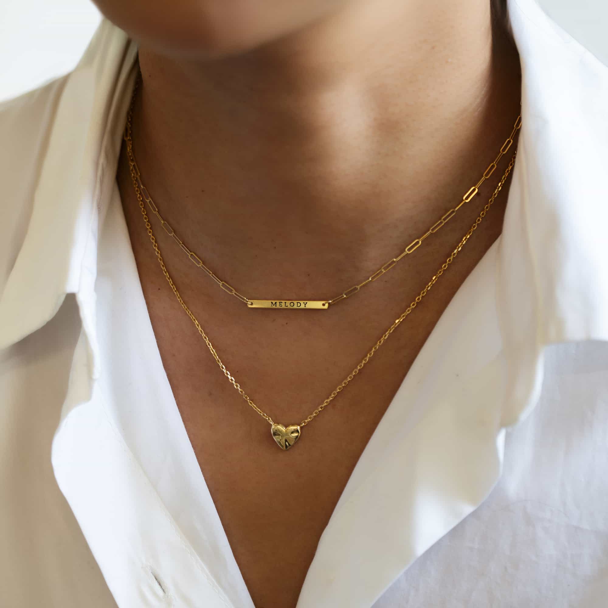Gold-filled thin bar necklace engraved with a name, layered with a gold heart pendant on a model