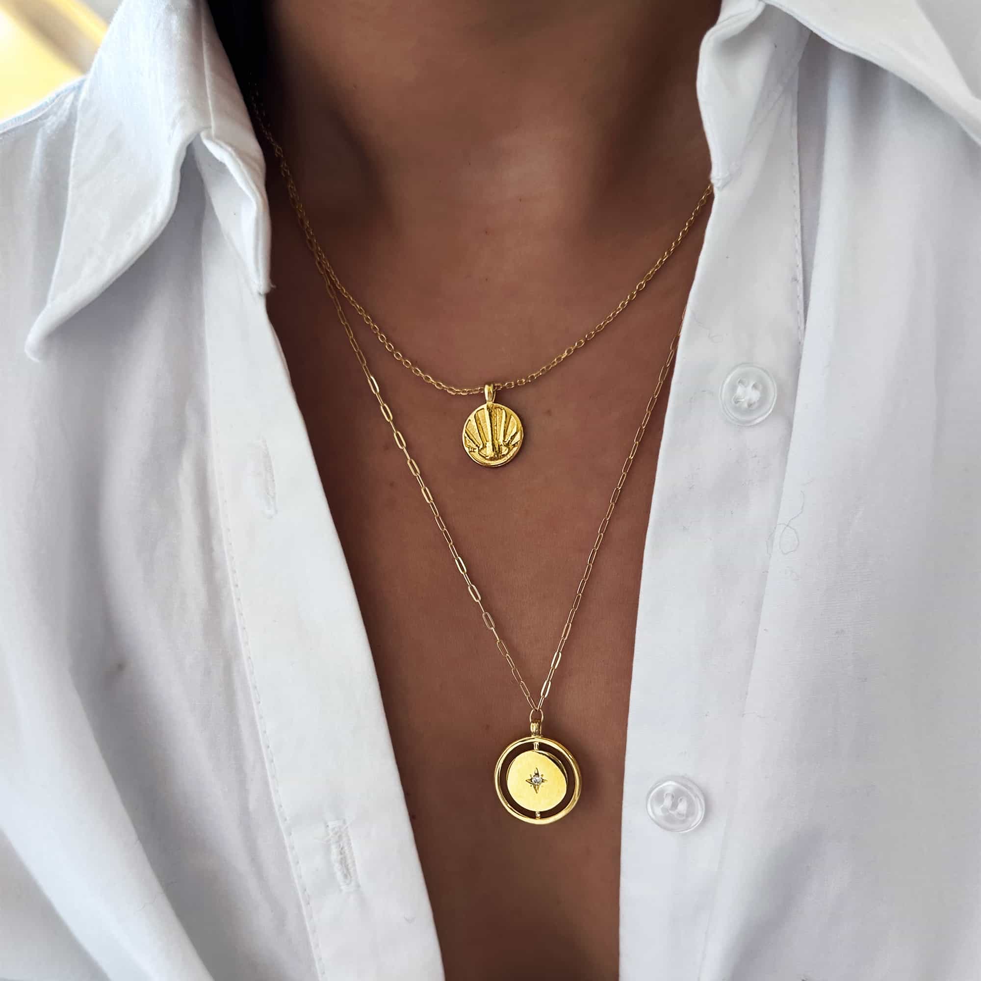 Two gold disc pendant necklaces, one shell motif and one with a clear stone, on a model's neckline