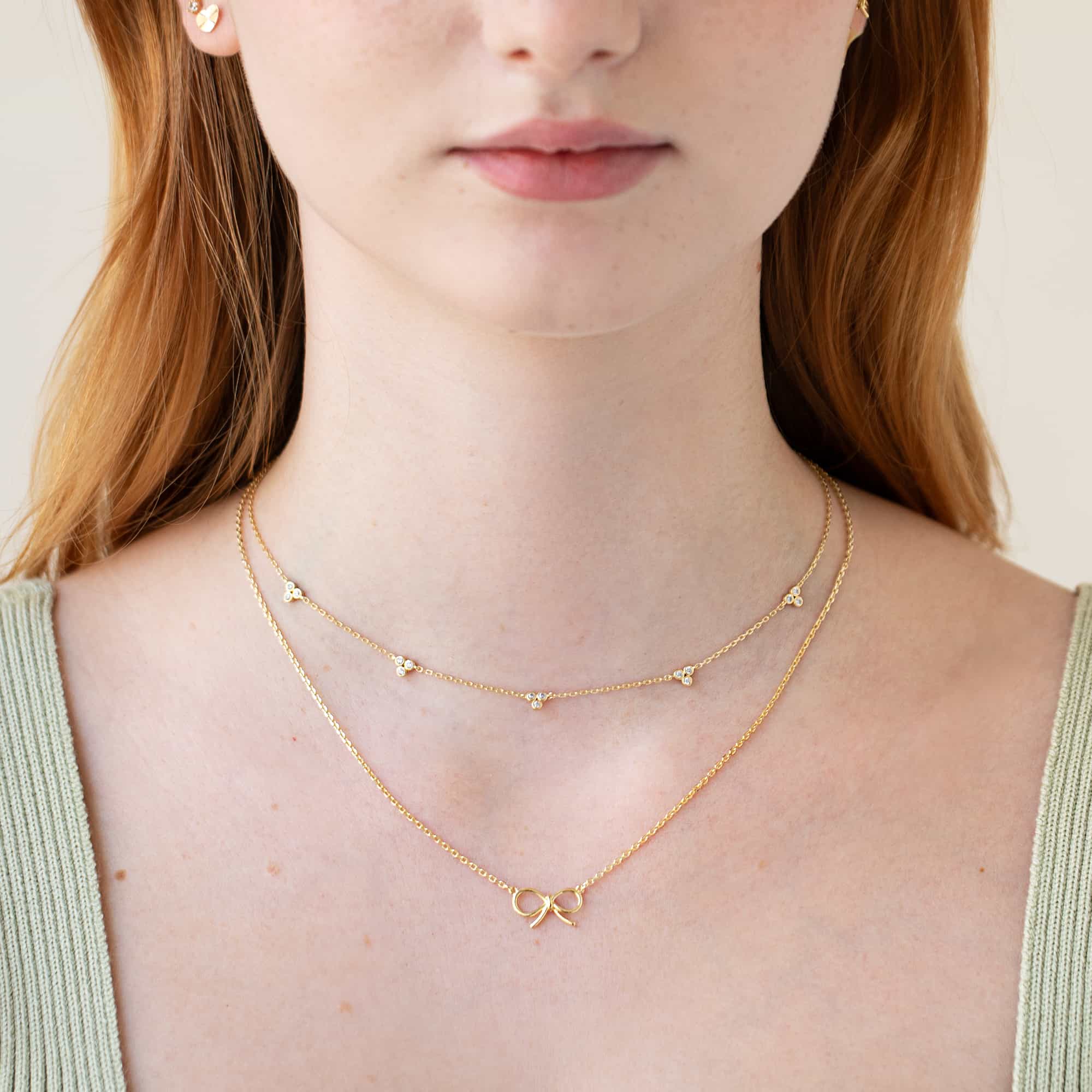 Layered gold necklaces, a clear-stone clover station chain and a bow pendant, shown on a model's neckline