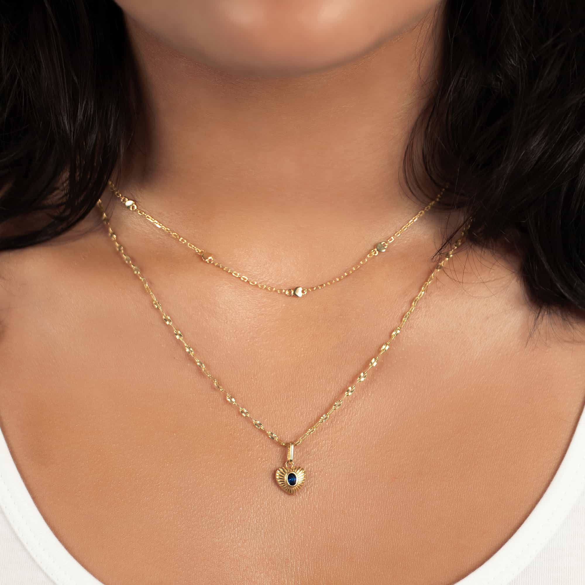 Gold heart pendant with a blue sapphire on a chain, shown layered on a model's neckline