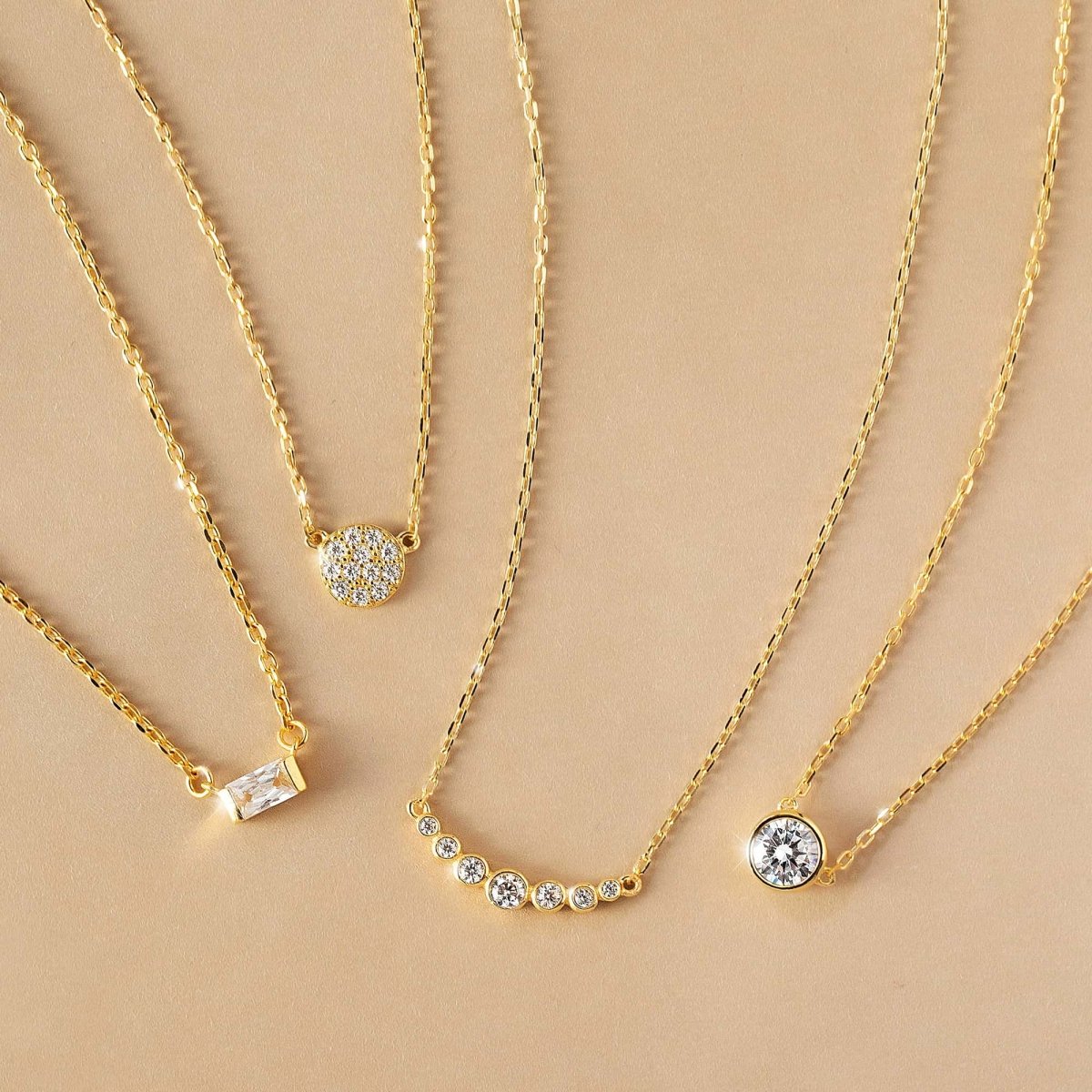Gold vermeil curved bar necklace with graduated round clear stones, shown with other pendant styles on a cream background