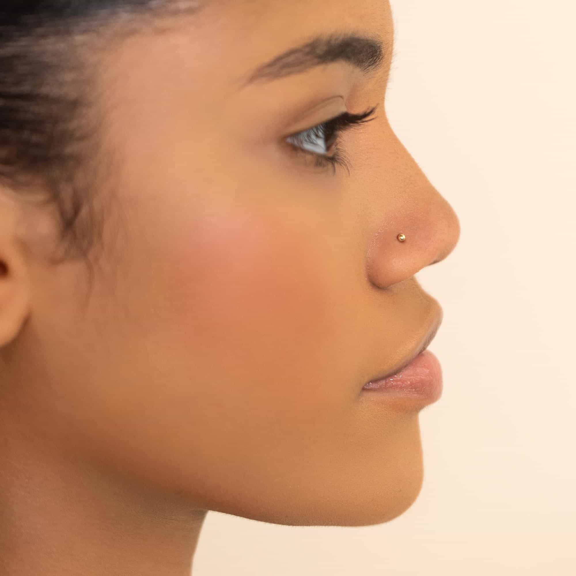 14K gold ball nose stud worn on a model's nostril, side profile against a cream background
