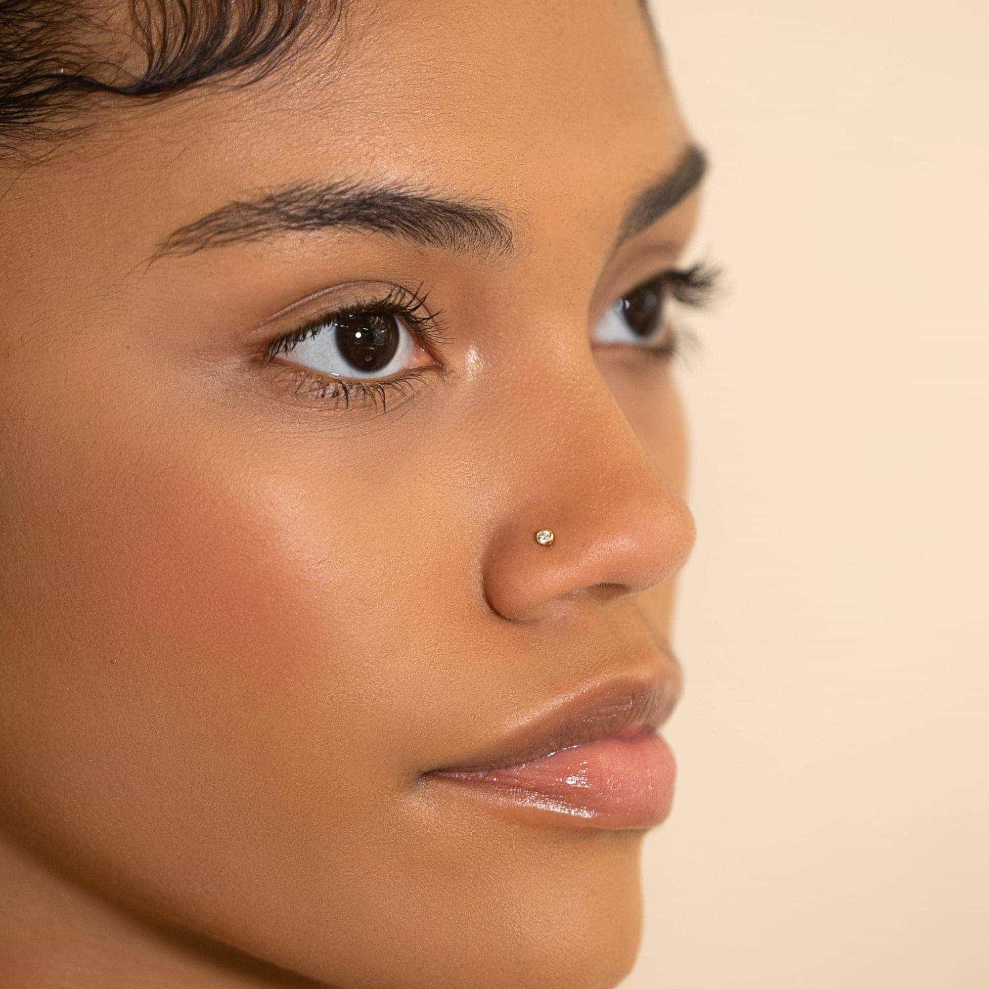14K gold bezel nose stud with a clear white zirconia, shown on a model's nose in profile