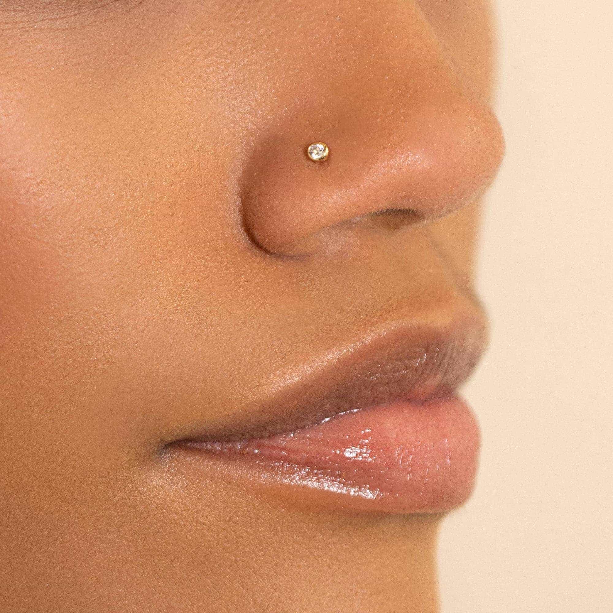 14K gold bezel nose stud with a clear white zirconia, shown on a model's nostril