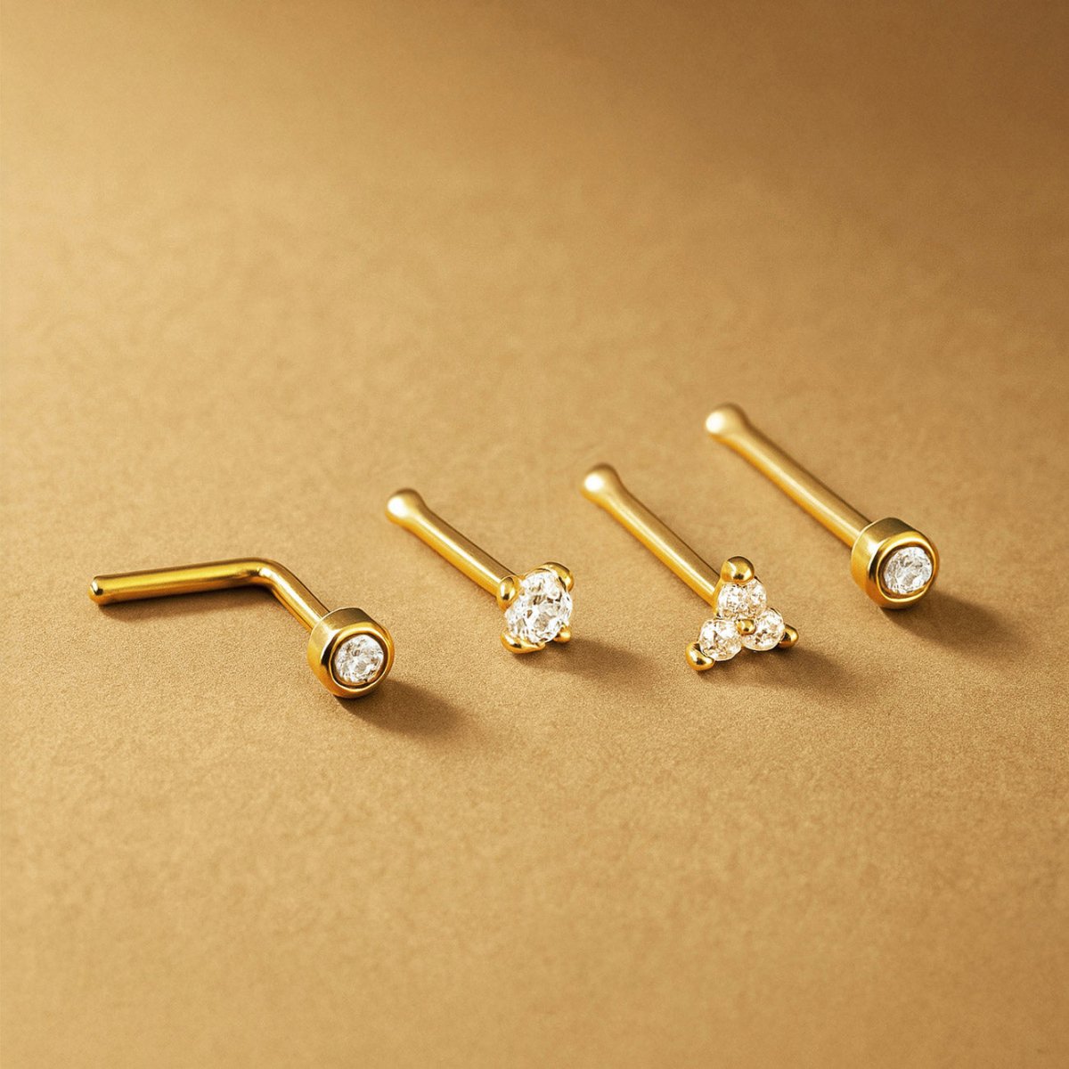 Four gold nose studs with clear stones in bezel, prong, trio and L-bend styles, on a tan surface