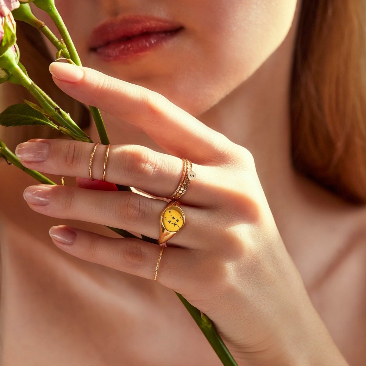 Gold-filled disc and heart initial rings stacked on a model's finger, shown with flowers