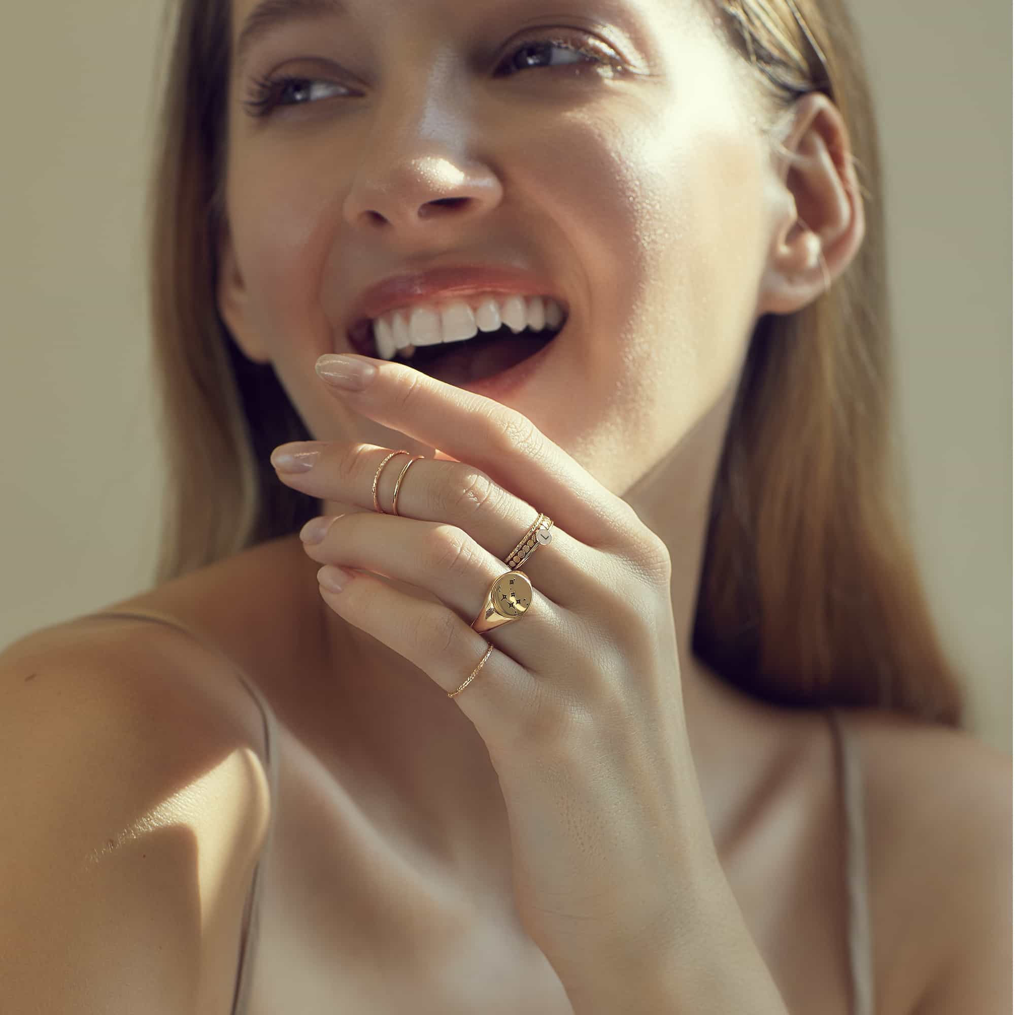 Stacked gold thin band rings and a round signet ring on a smiling model's hand near her chin