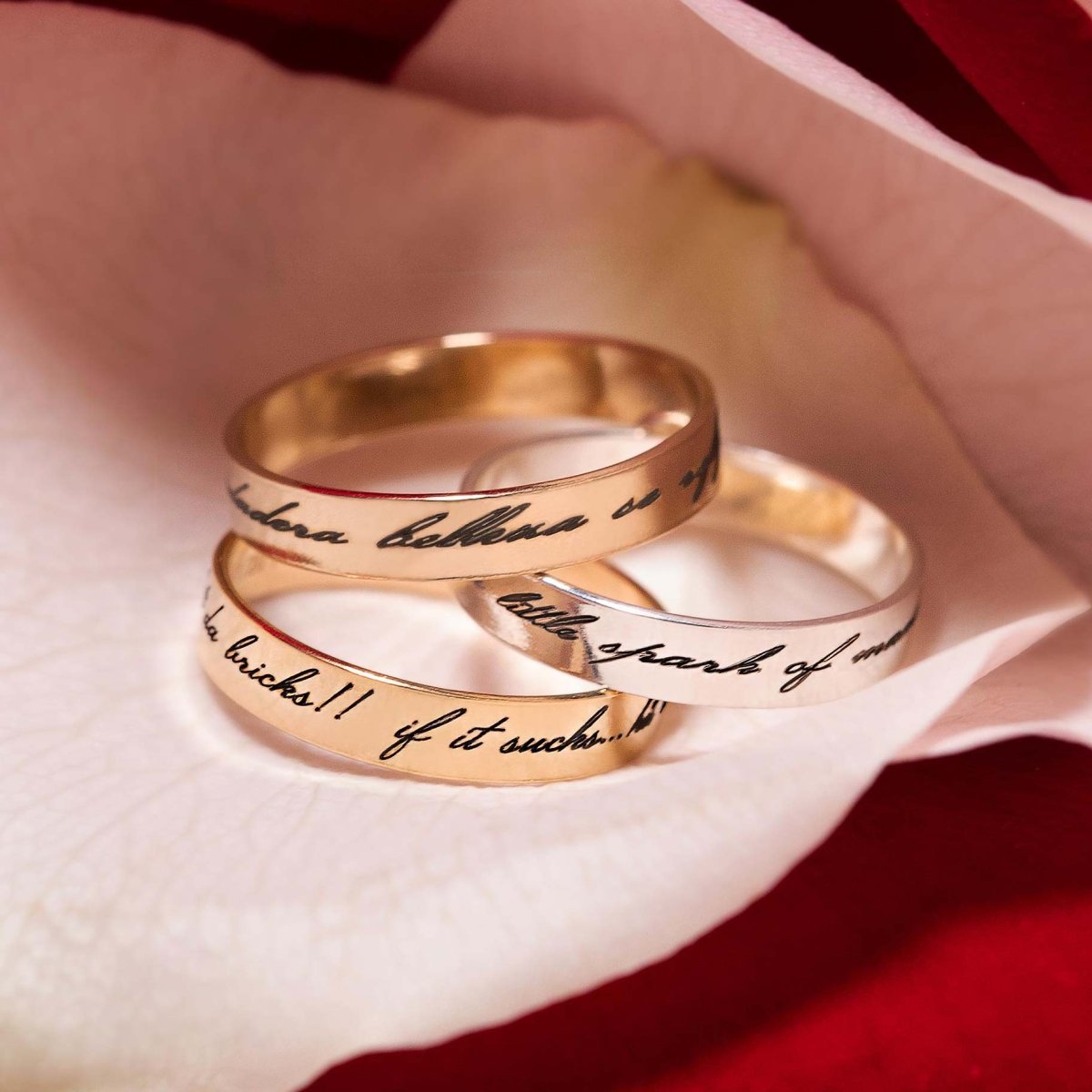 Stacked gold-filled and silver engraved band rings with script messages, resting on a rose petal