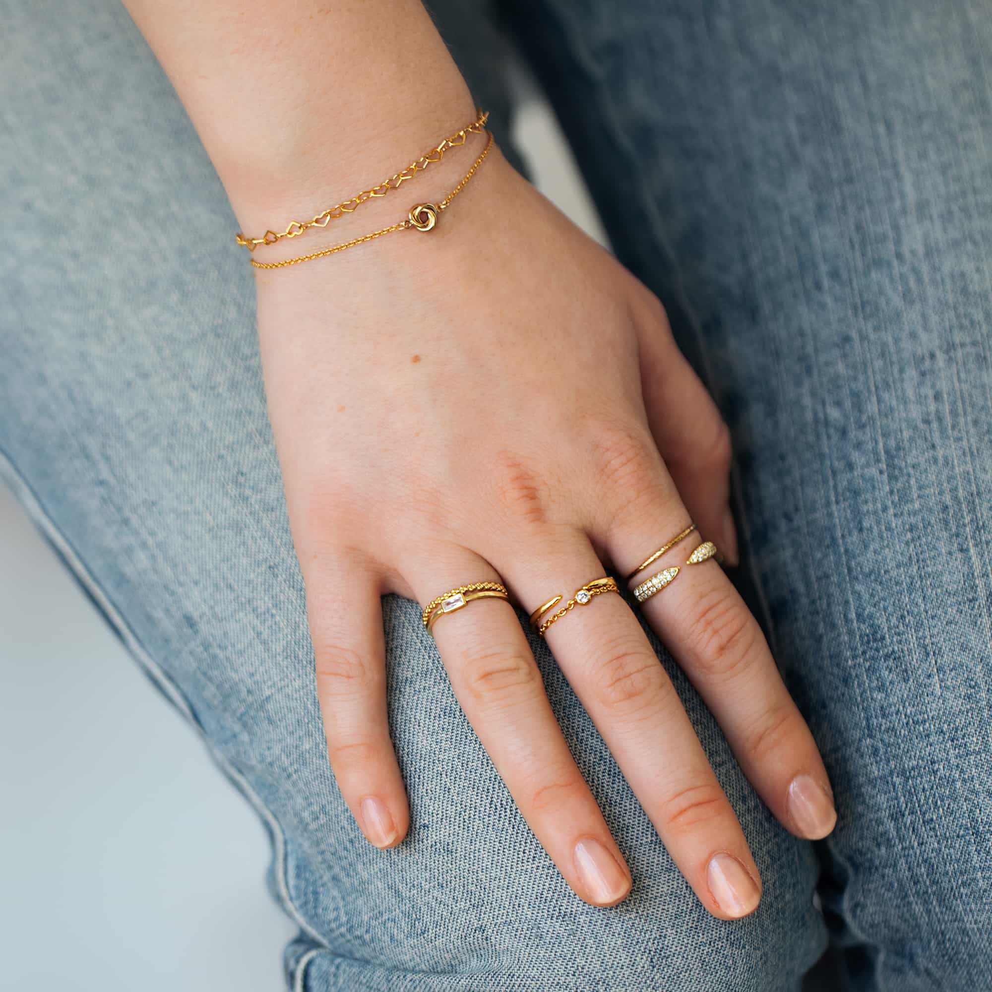 Gold-filled tiny love knot chain bracelet shown layered on a model's wrist with stacked rings
