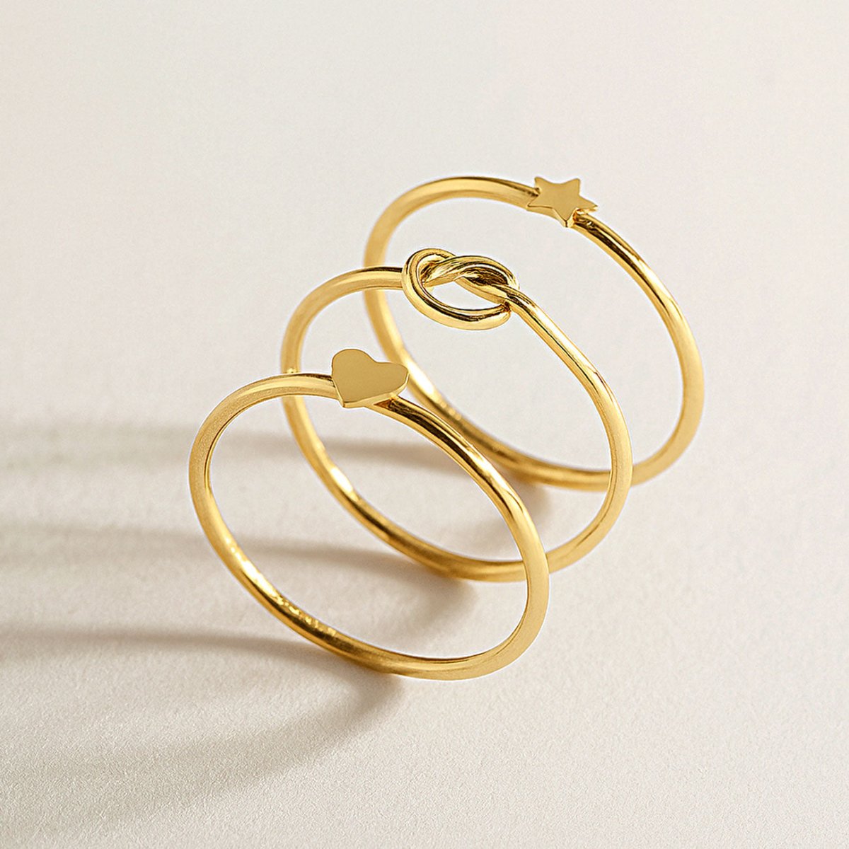 Three gold thin stacking rings with star, knot and heart accents on a cream background