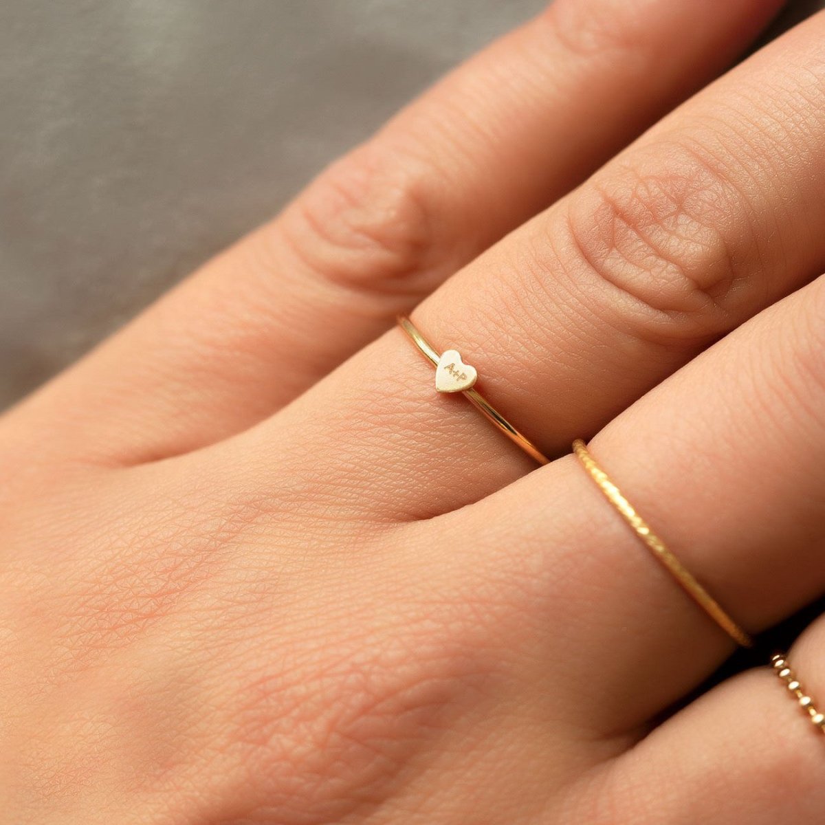 Gold-filled tiny heart ring engraved with initials, close-up on a model's finger beside a thin band