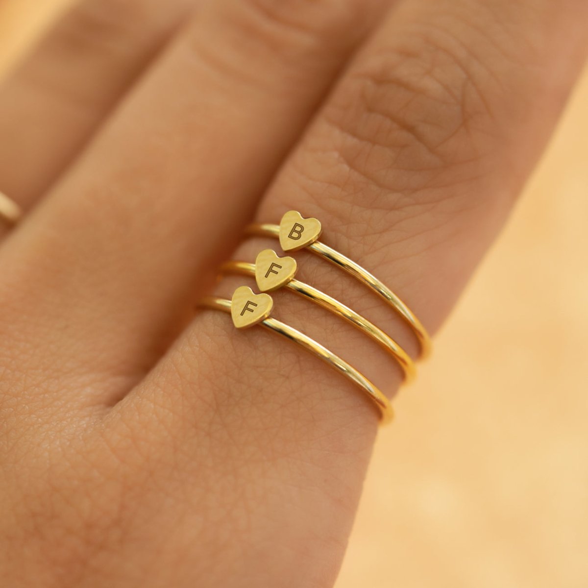 Gold-filled stacked heart rings, each engraved with an initial, shown on a model's finger