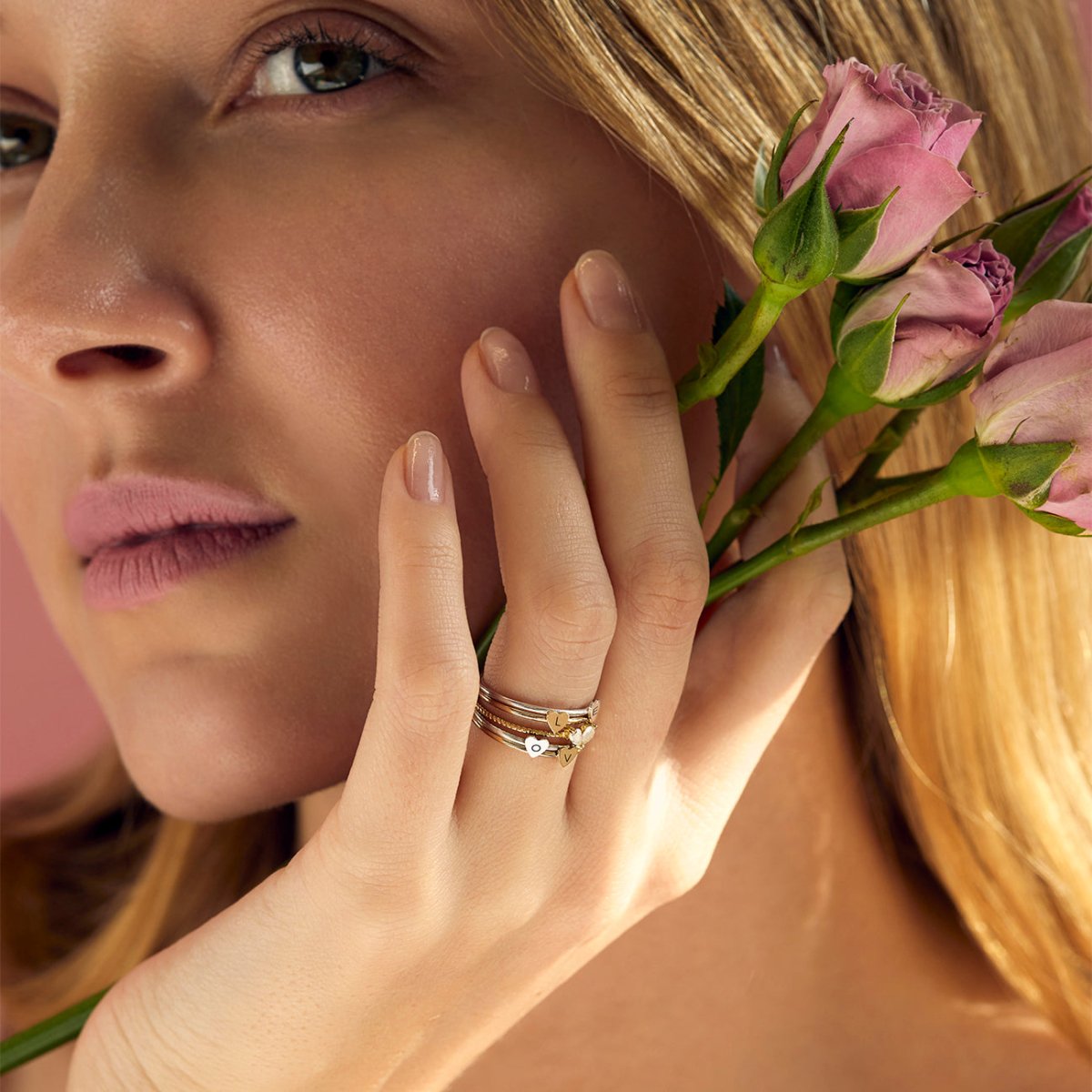 Gold-filled heart initial rings stacked on a model's finger, held beside pink roses