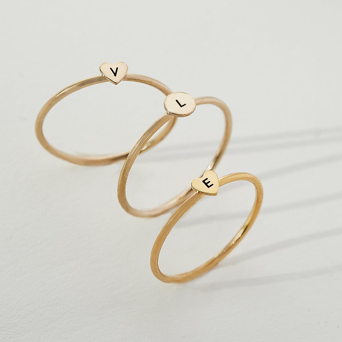Gold-filled stackable rings topped with heart and disc charms engraved with initials, on a cream background