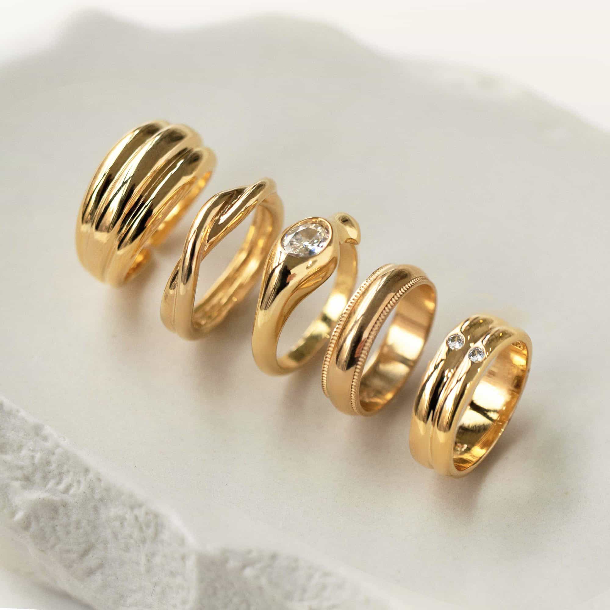 Row of five gold rings, including a domed band and a clear stone ring, on white marble