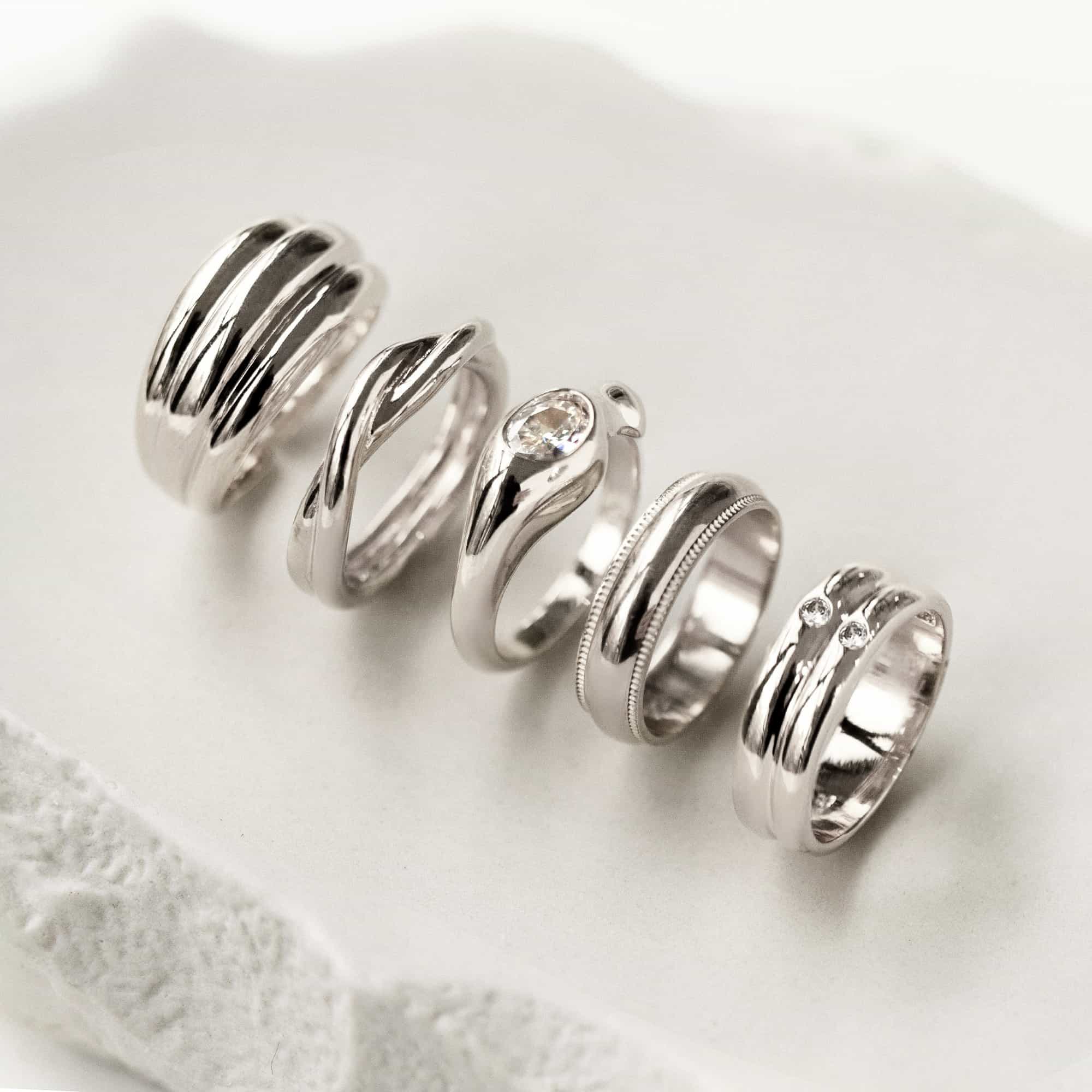 Five silver rings in varied band styles, including a clear-stone ring, on a cream surface