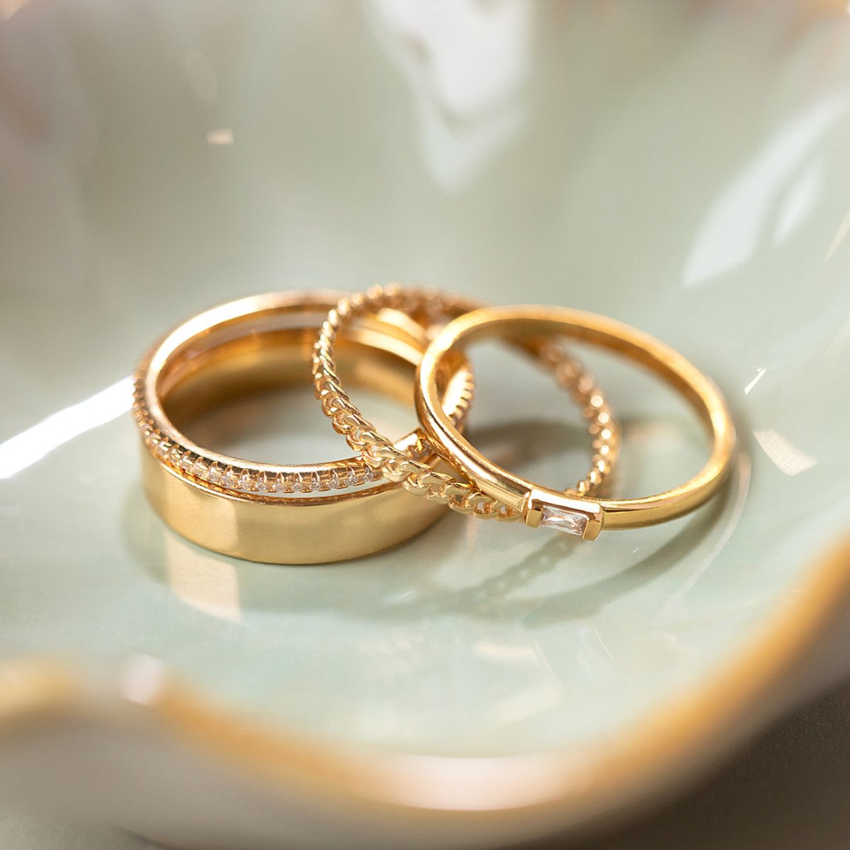 Gold ring set with a wide band, a curb-chain ring and a clear-stone band, in a cream dish