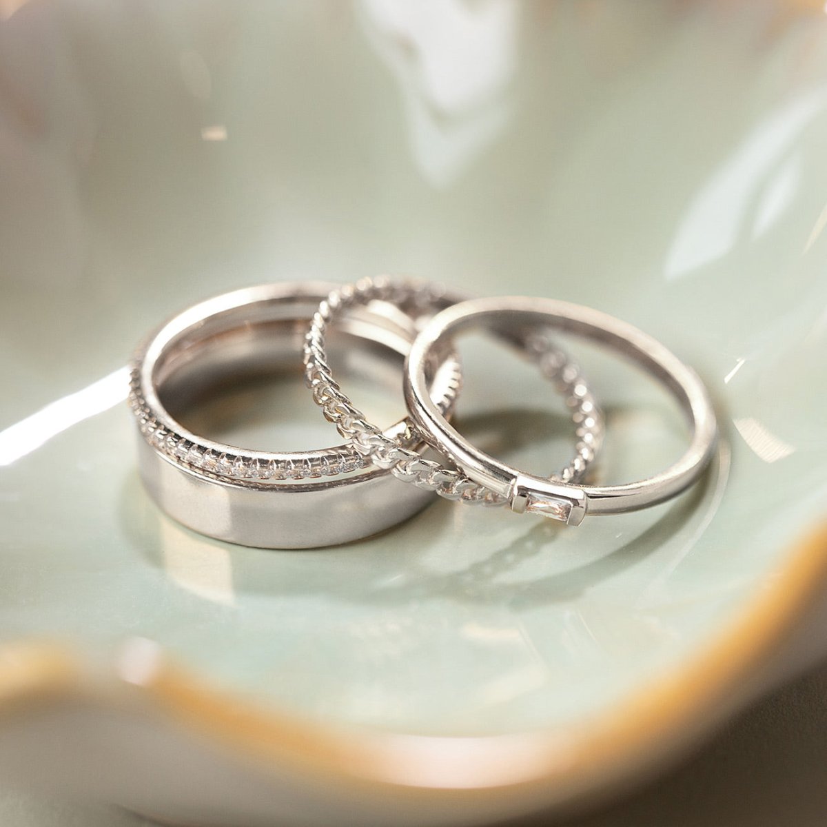 Silver ring set with a wide band, a curb-chain ring and a clear-stone band, in a cream dish