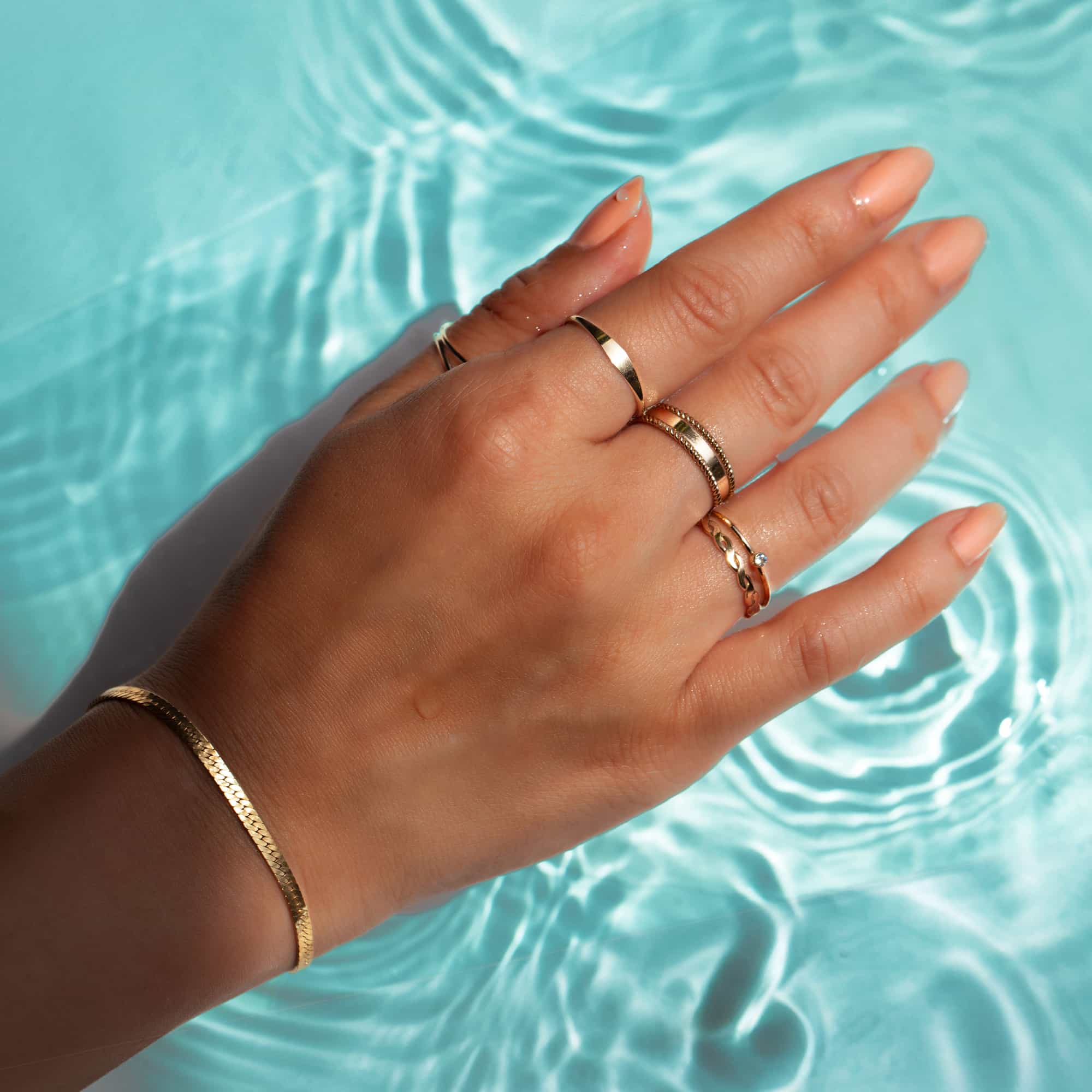 Stacked gold band and rope rings on a hand resting above rippling blue water