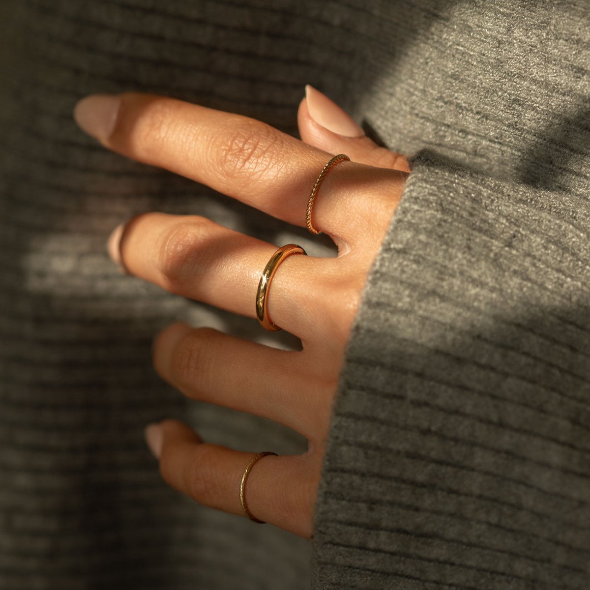 14K gold thin band rings, plain and twisted, shown stacked on a model's fingers against a grey sweater
