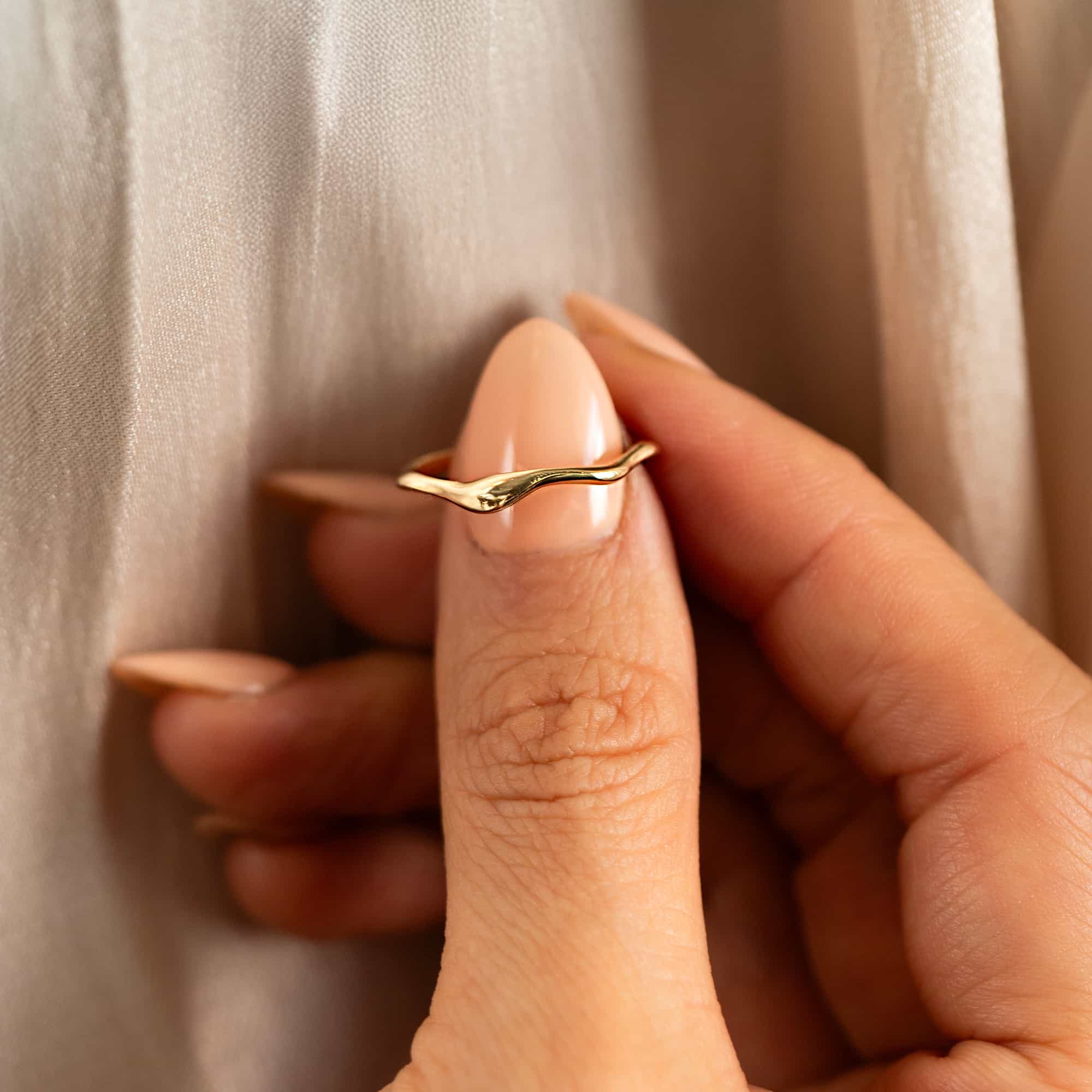14K gold dainty wave stacking ring held between fingertips against cream fabric
