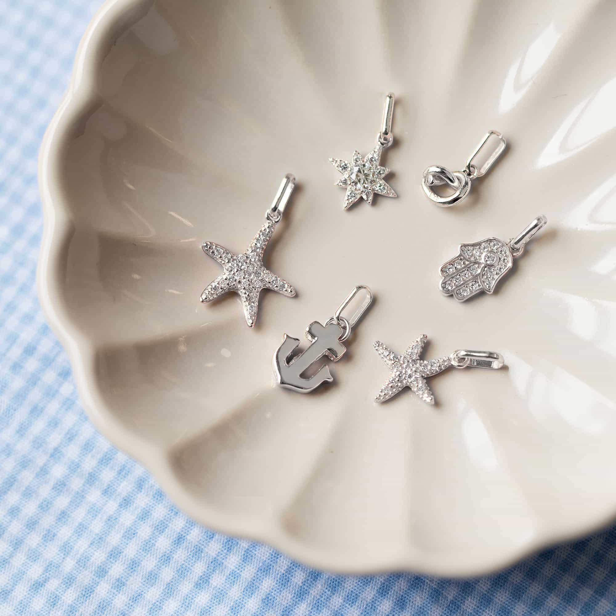 Assorted silver charms including starfish, star, hamsa and anchor in a shell dish on blue gingham