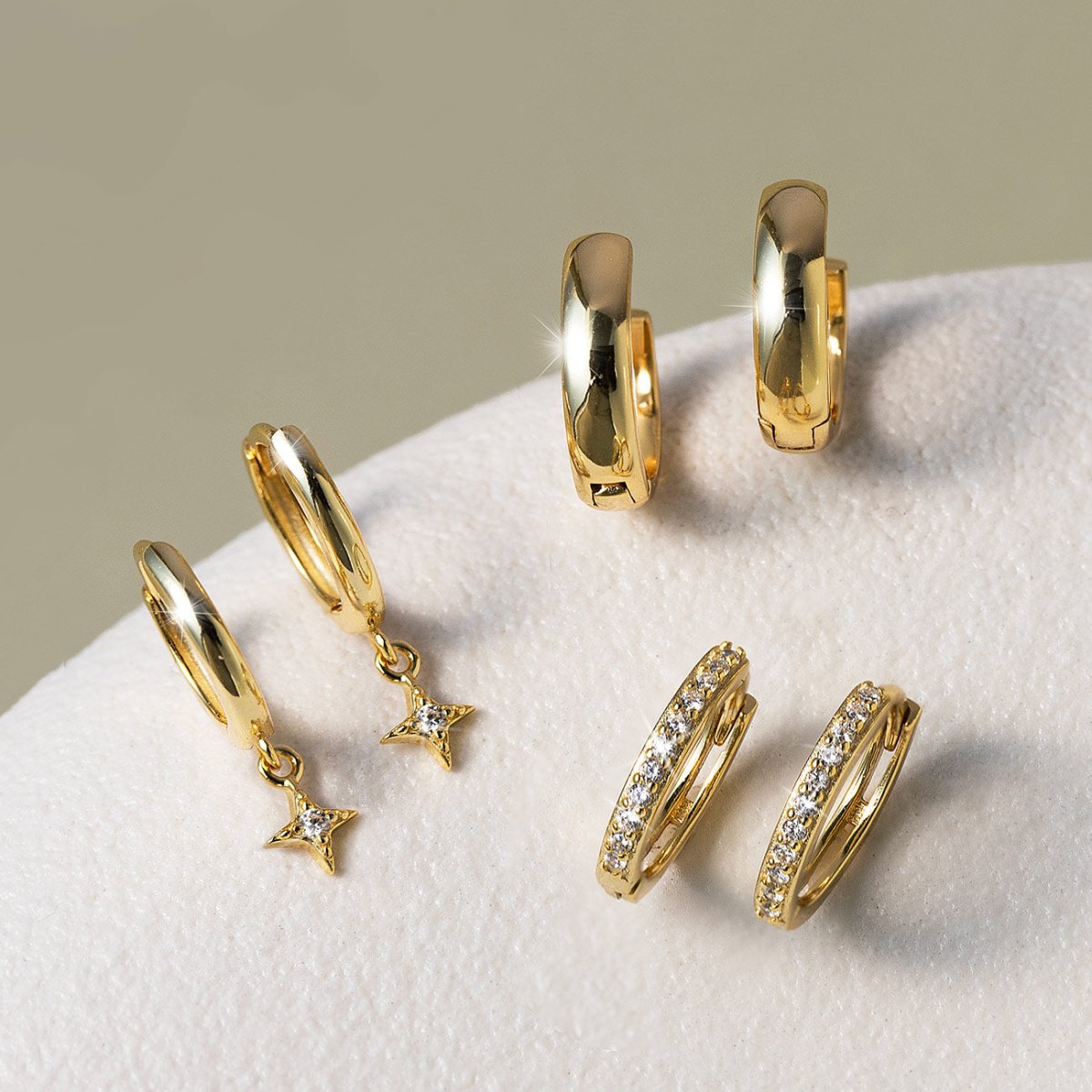 14K gold pavé huggie hoops and plain huggies with starburst charms set with clear stones, on a cream background