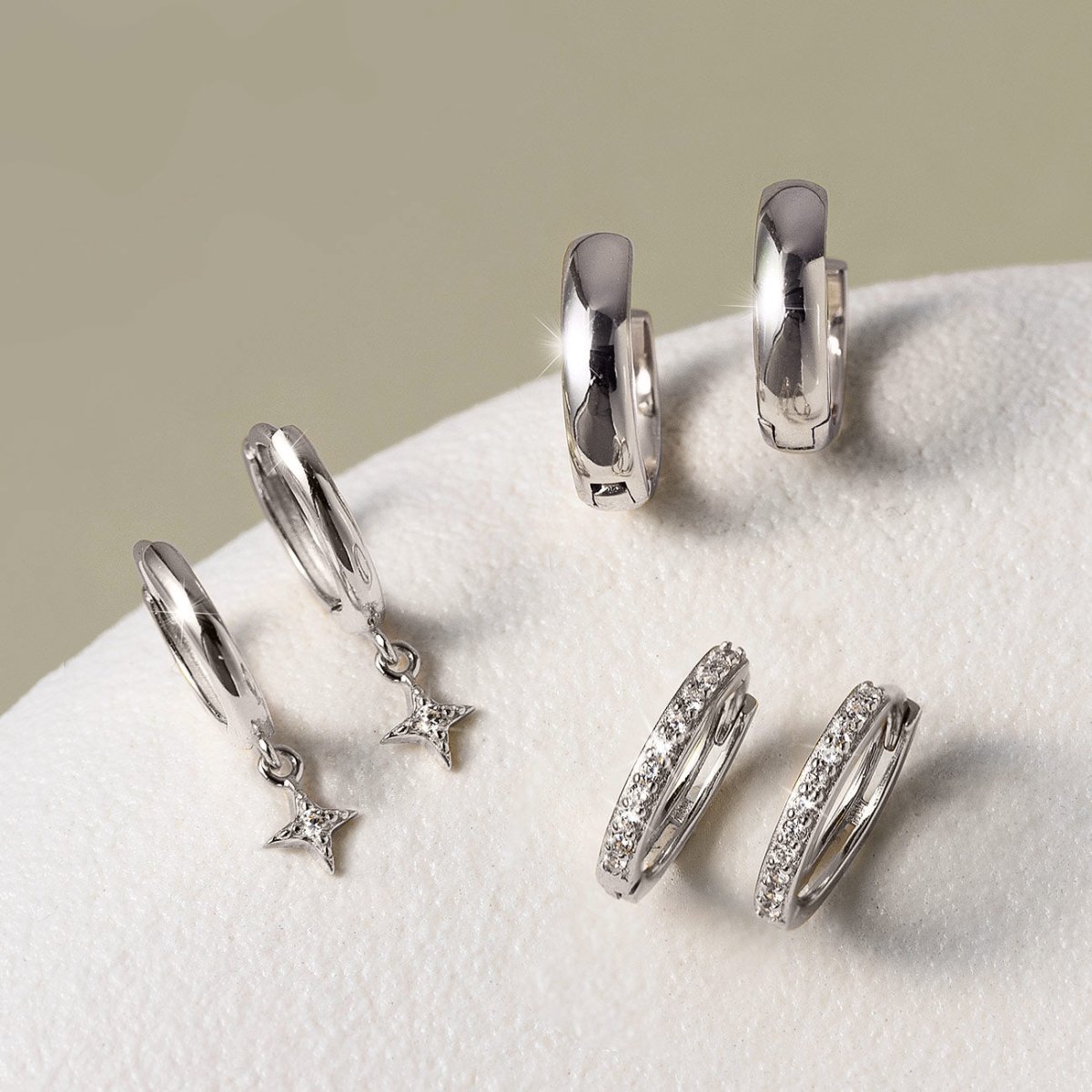 Three pairs of silver huggie hoops, including plain, pavé and star-drop styles, on a cream background