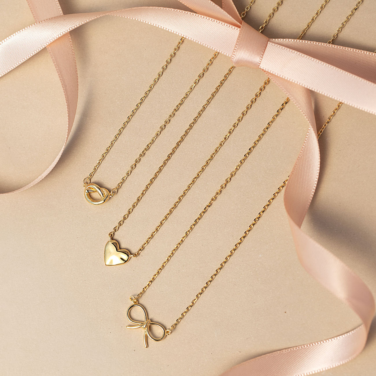 Three gold necklaces from AMYO with heart, knot, and bow pendants on a beige surface with pink ribbons perfect for gifting.